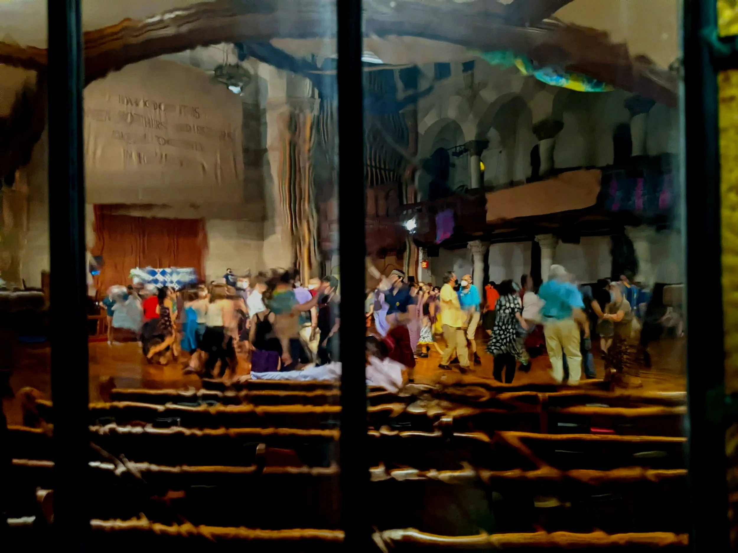 People dancing inside a church with wooden pews and a balcony, seen through a glass window.