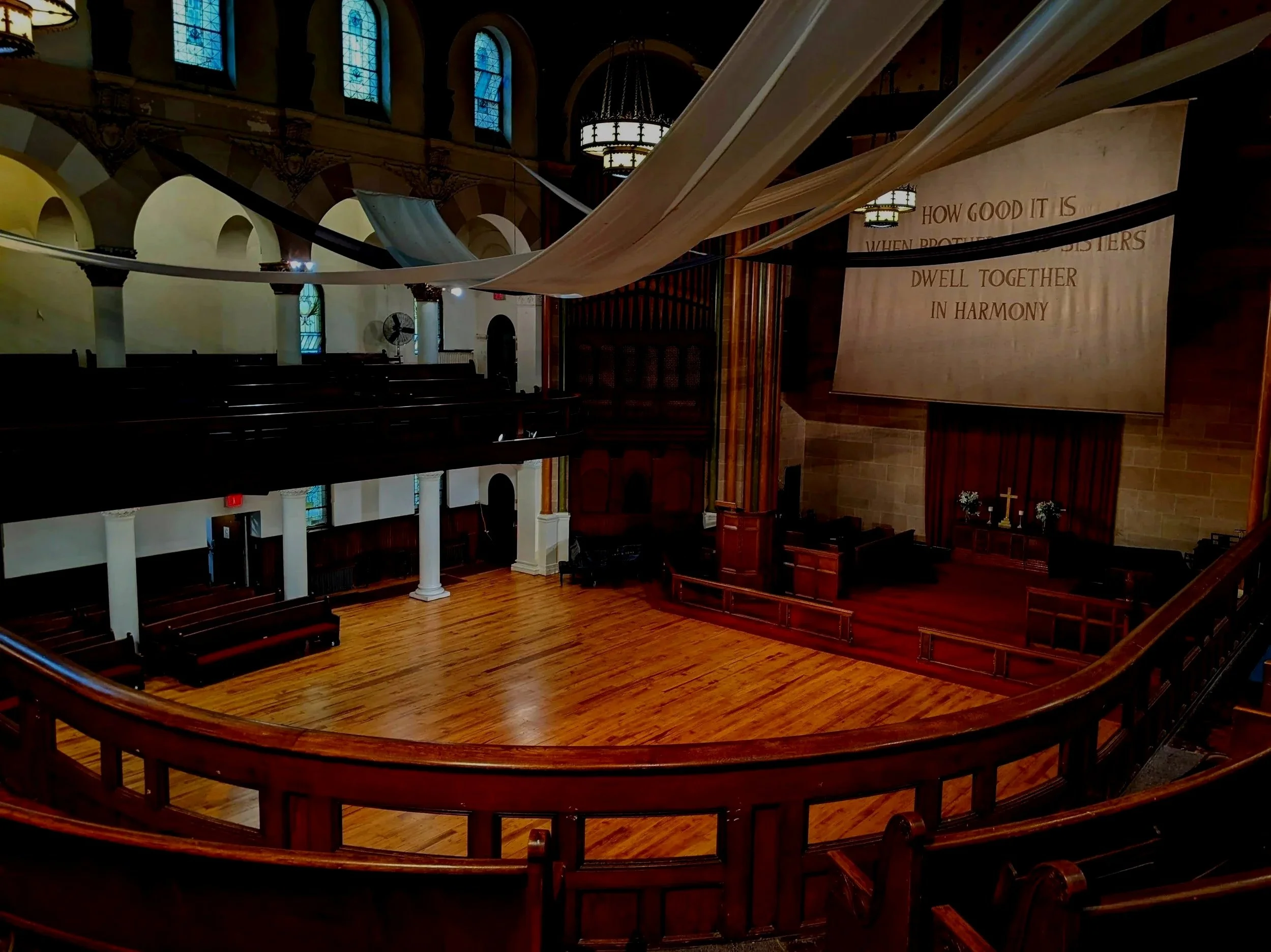 Empty church sanctuary with wooden pews, elevated balcony, stained glass windows, hanging lights, a large banner with the quote 'How good it is when brothers dwell together in harmony', and a small altar with a cross and flowers.