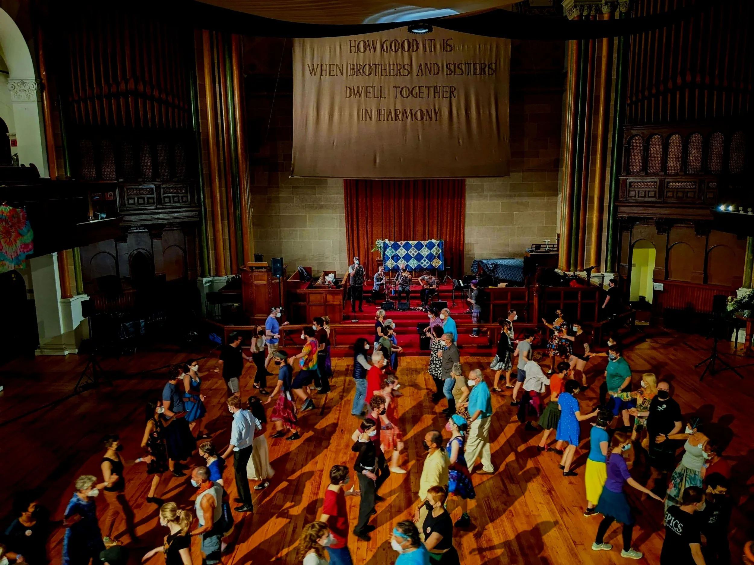 People dancing in a large hall with wooden floors and an elevated stage at the front. The stage has a choir and a quilt hanging on the back wall. A large banner hangs above with the words: 'How good it is when brothers and sisters dwell together in harmony.'