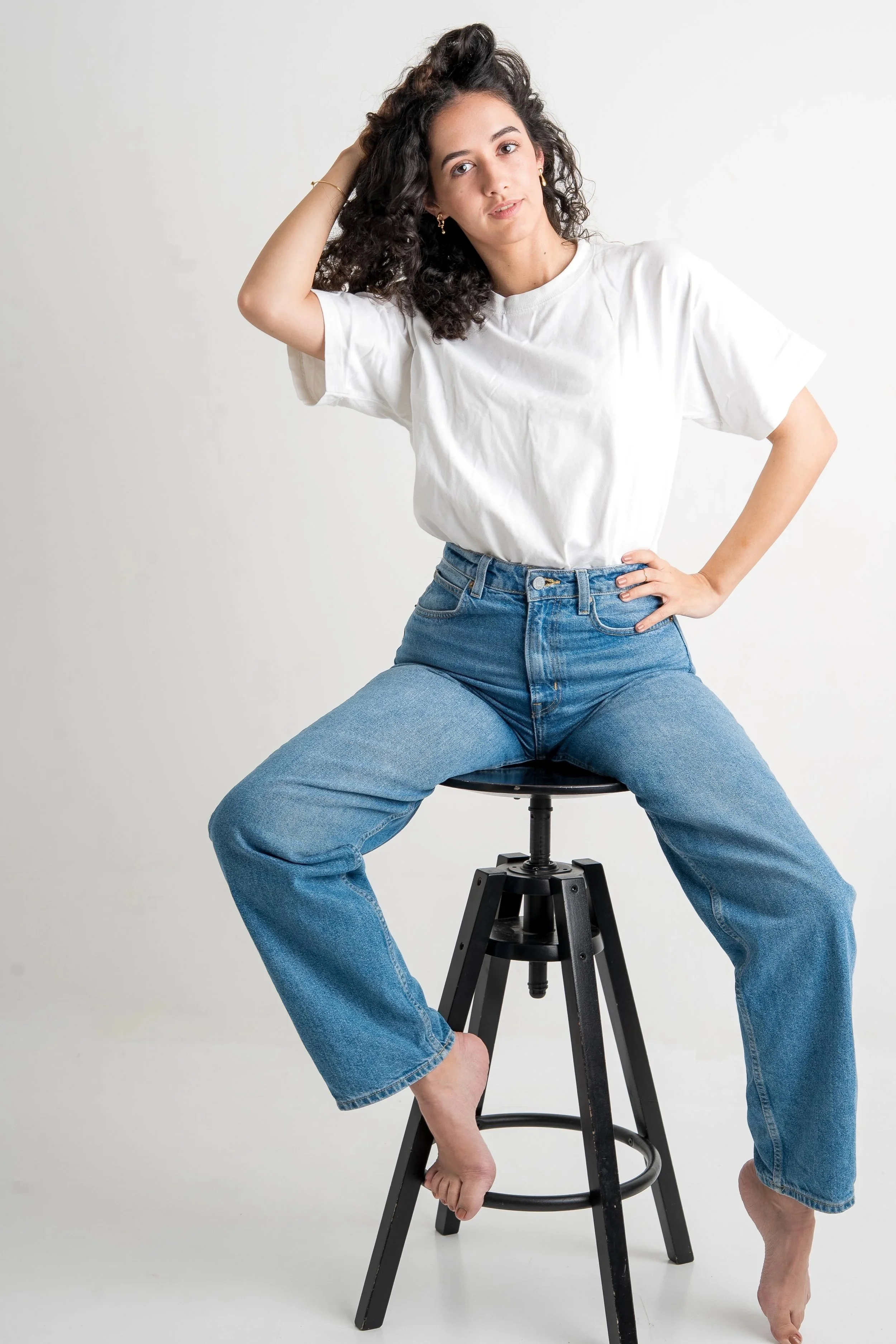 Junge Frau sitzt auf einem schwarzen Hocker, trägt weiße T-Shirt und blaue Jeans, hat lockiges braunes Haar, schaut in die Kamera, hebt eine Hand zum Kopf, die andere auf die Hüfte, Barfuß