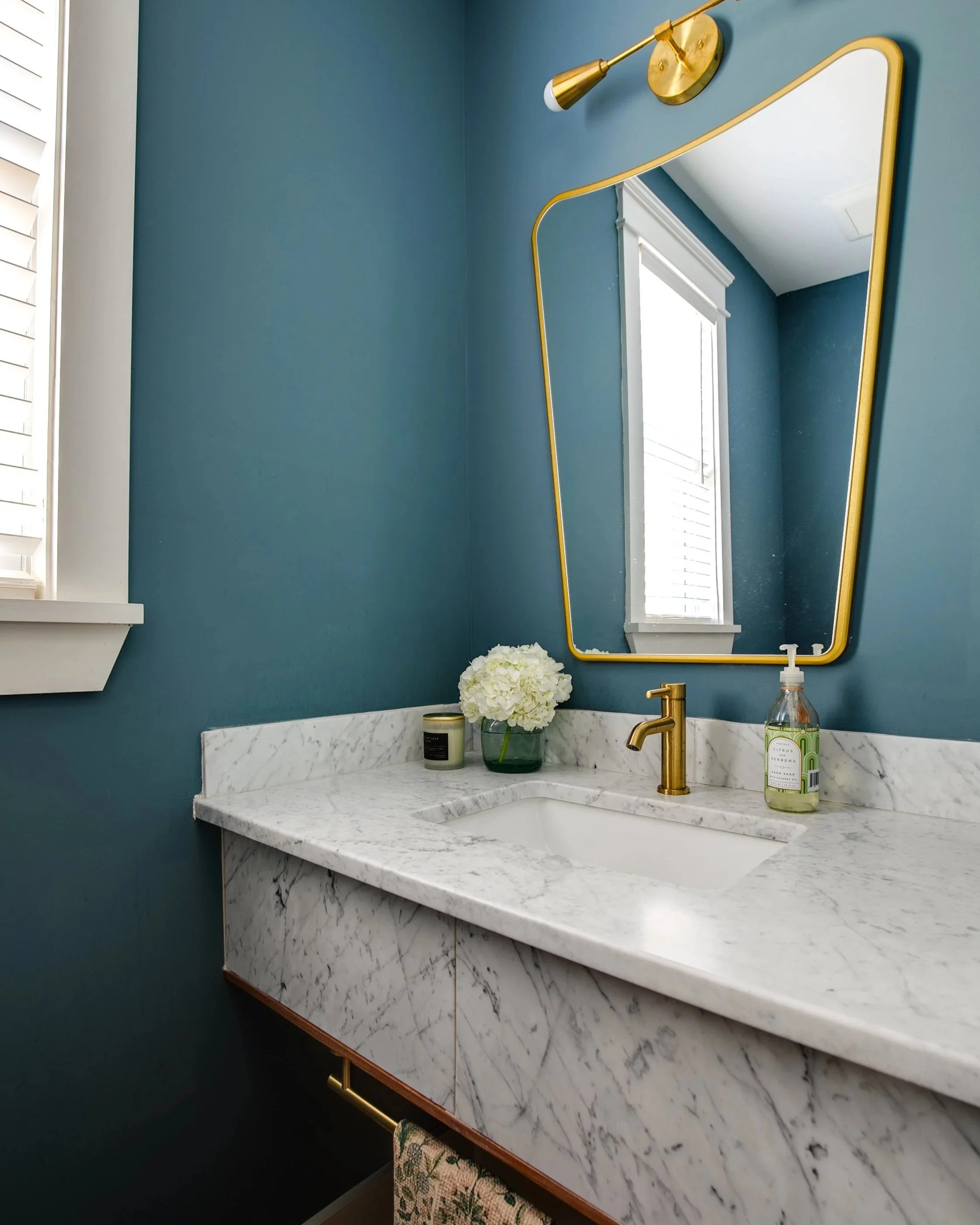 Custom bathroom design from Designs Delivered featuring deep teal walls, a marble vanity with brass fixtures, and a unique asymmetrical gold mirror, showcasing the personalized results available in the design customization service