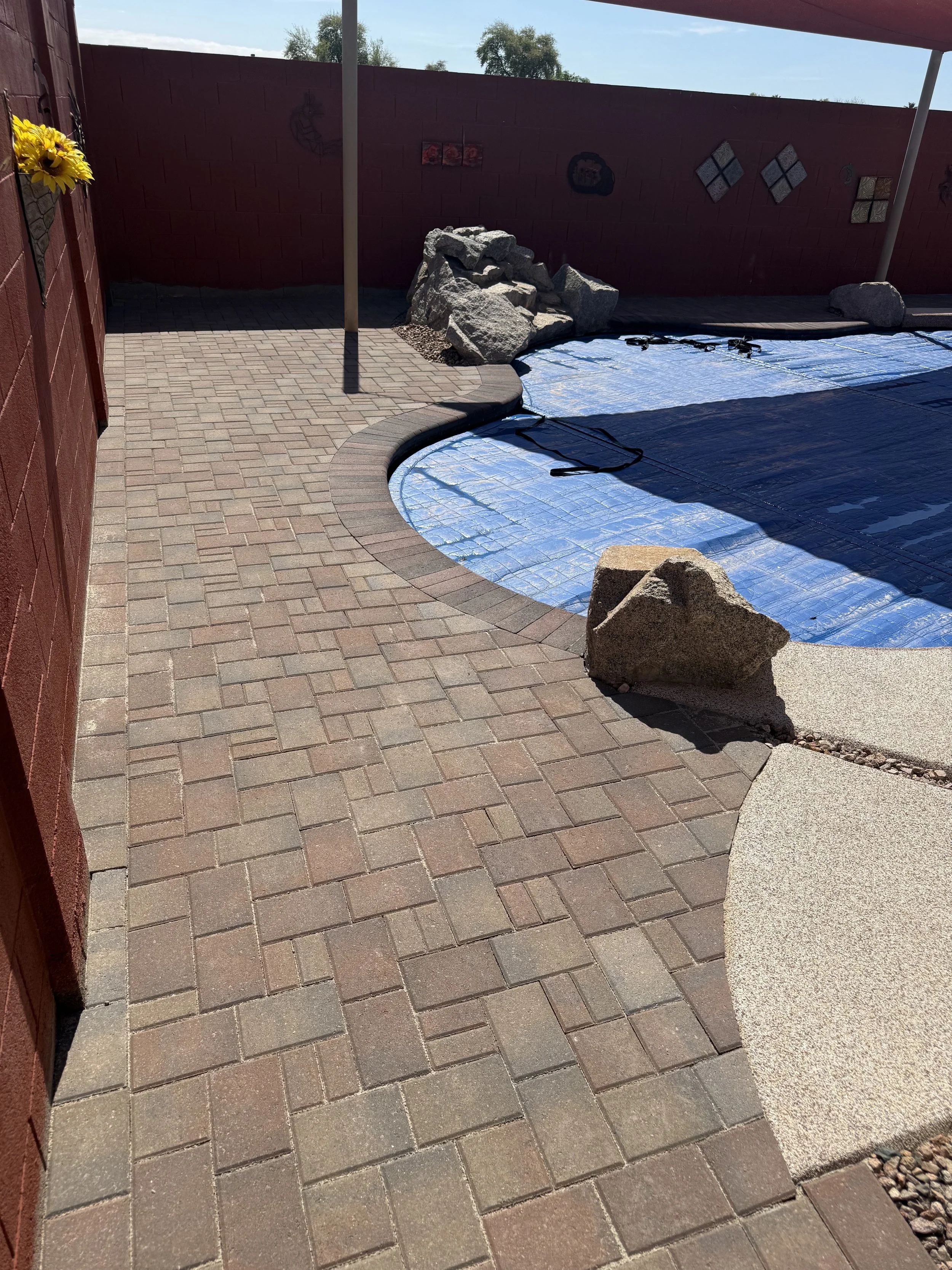 A paved driveway with interlocking bricks leading to a gate, adjacent to a garden bed with small shrubs and gravel, part of a residential property.
