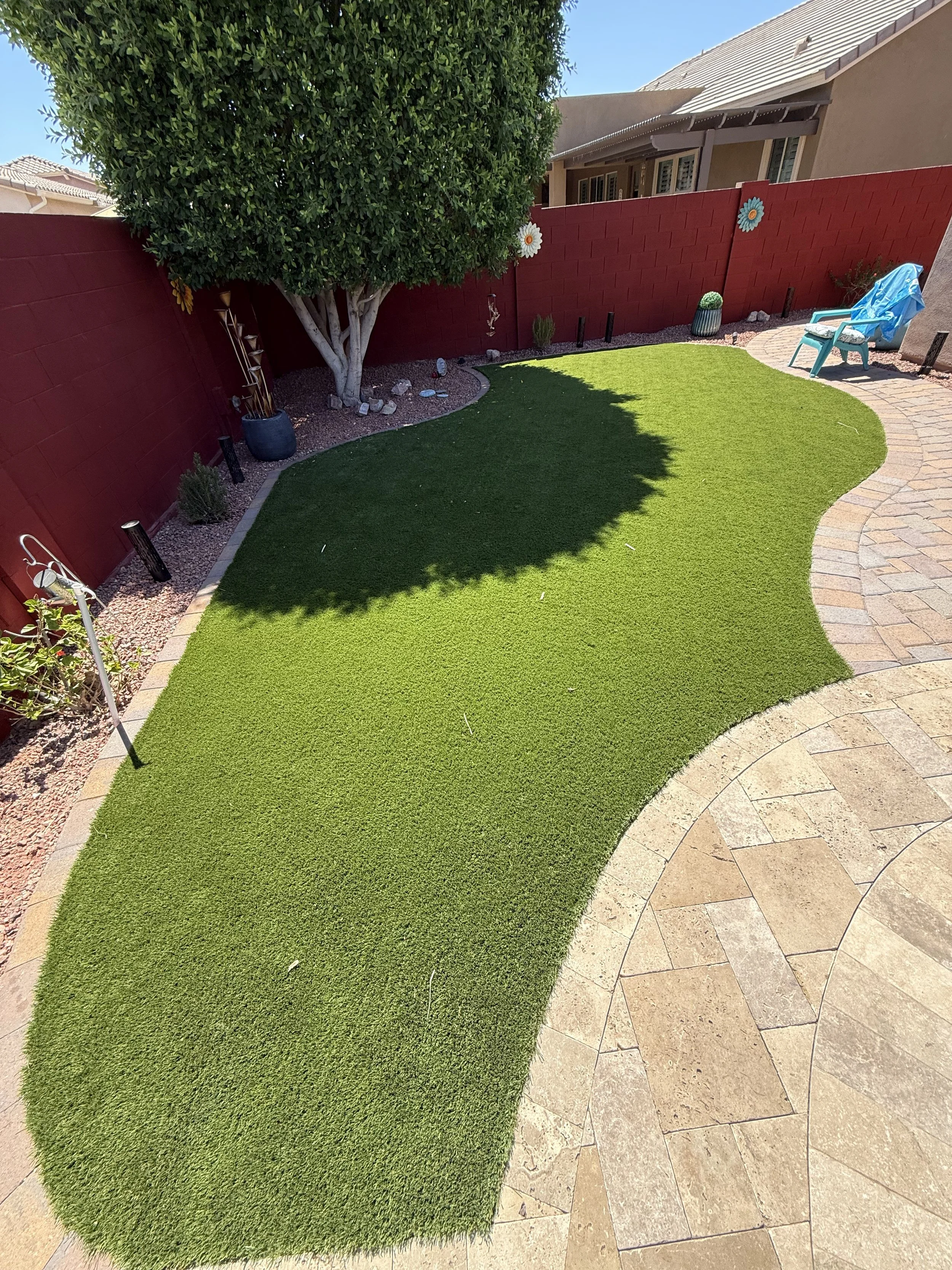 A backyard with a brick patio, artificial grass, palm trees, and a desert landscape with rocks, surrounded by a beige block wall under a blue sky with scattered clouds.