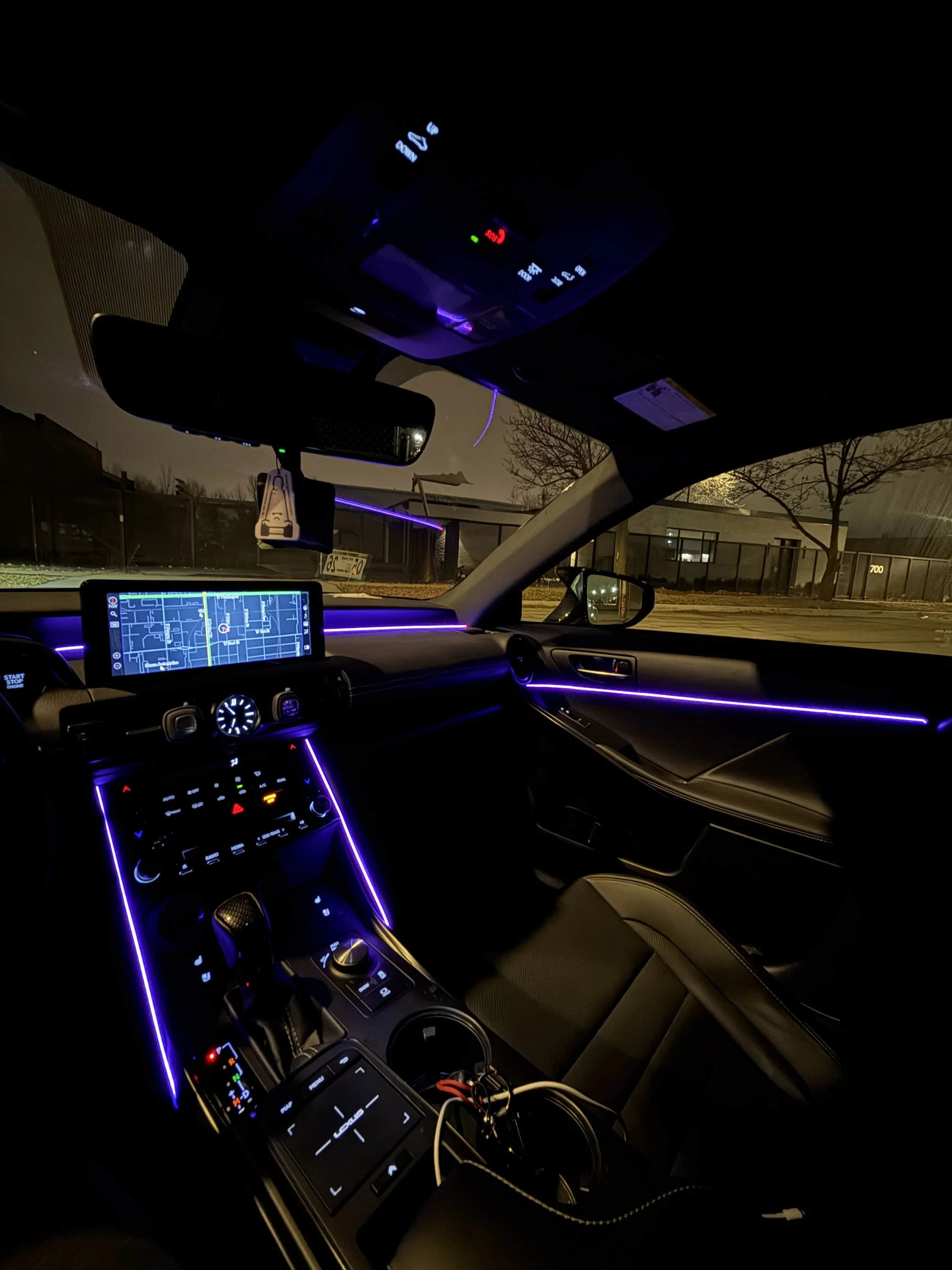 Interior of a car at night with illuminated purple LED accents, a navigation screen showing a map, and various controls and a rearview mirror.