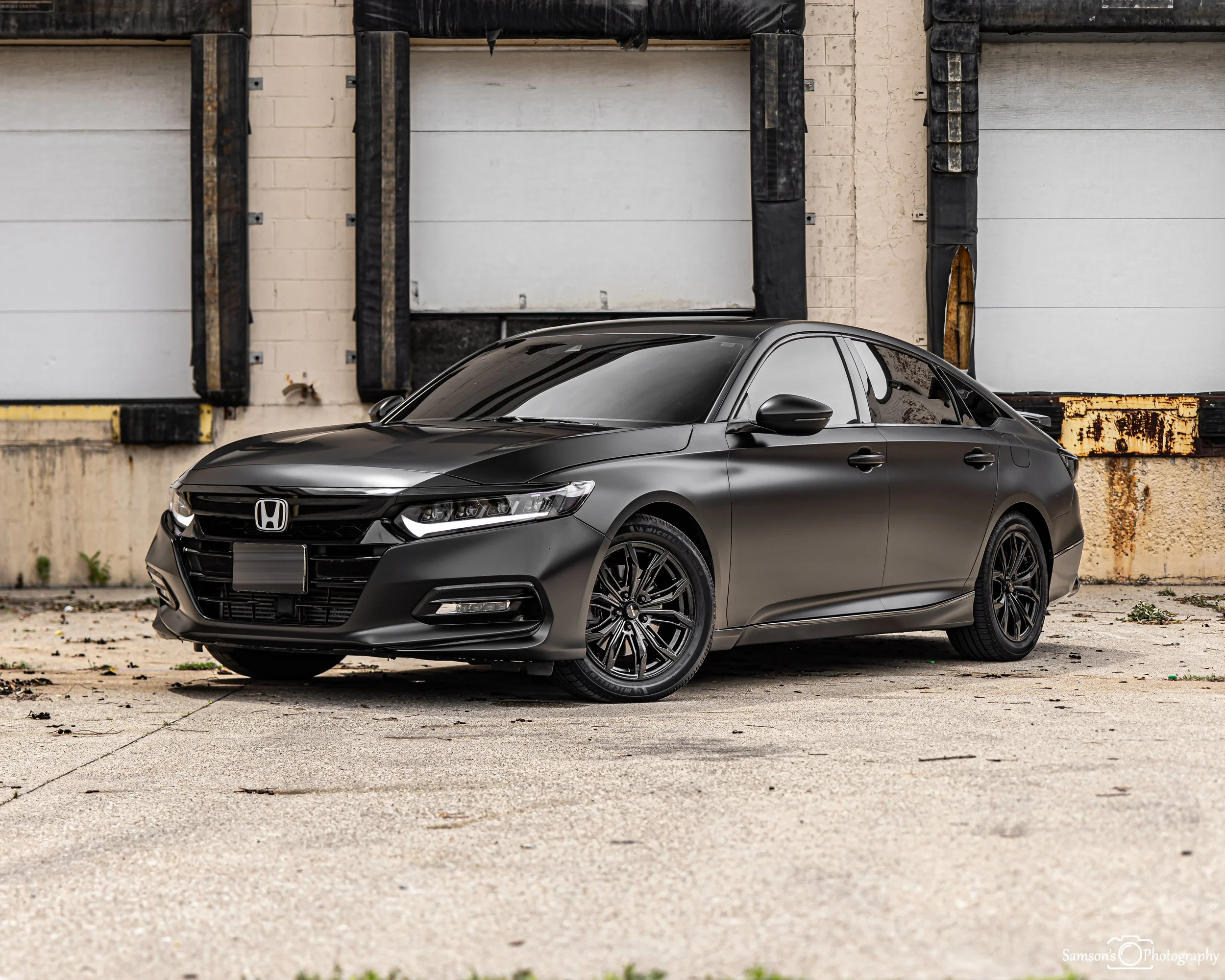 A black modern Honda car parked in an empty outdoor lot with a beige brick wall and garage door in the background.