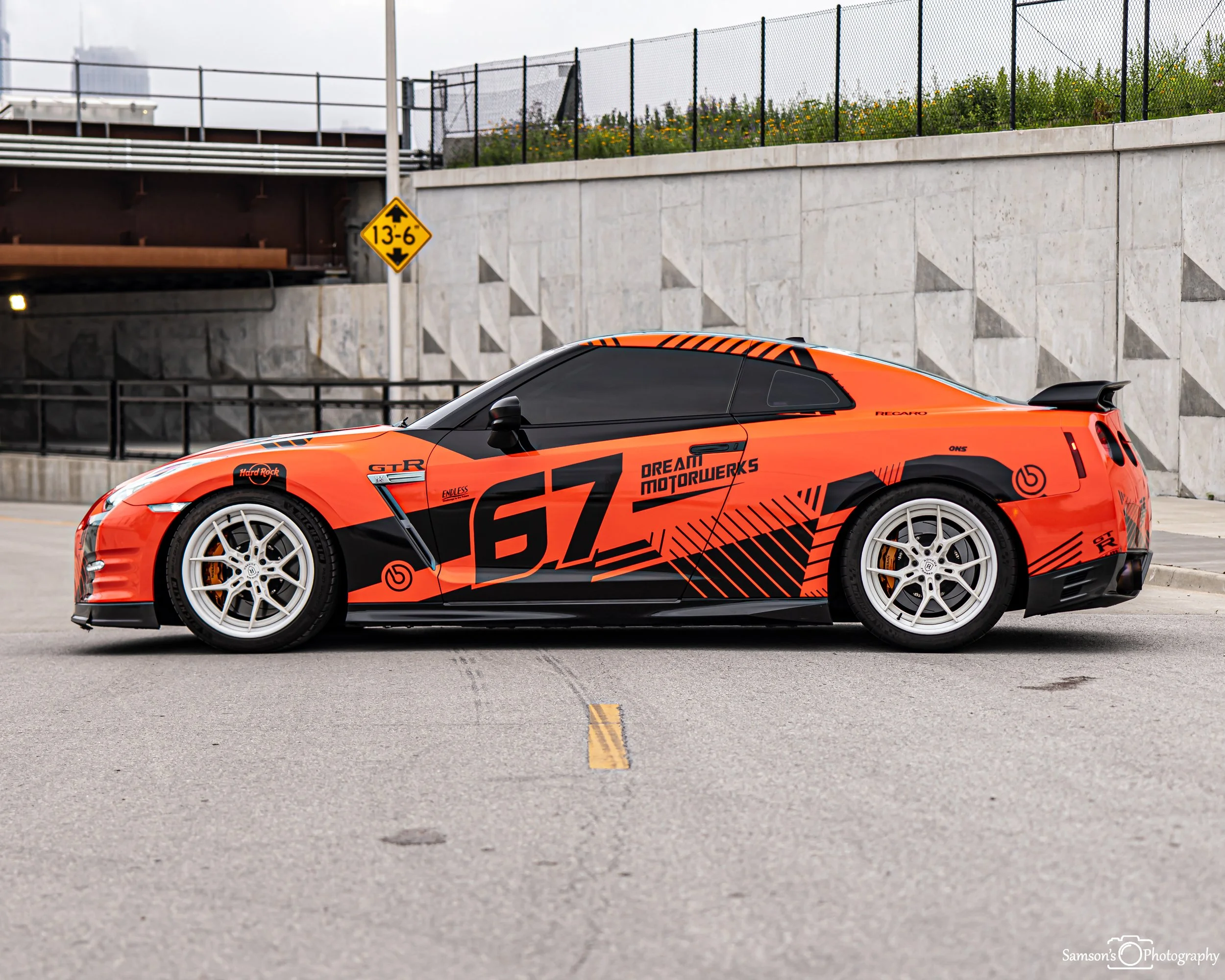 Orange race car with black graphics and sponsorship decals, parked in an outdoor parking lot.