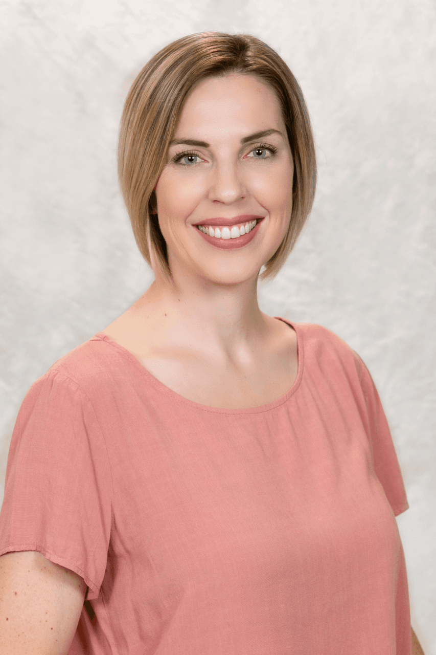 Smiling person with short blonde hair wearing a pink shirt against a light gray background.