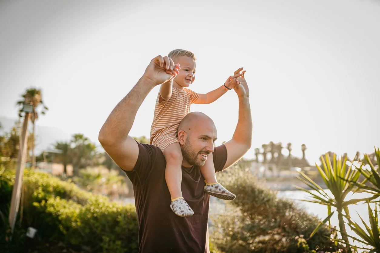 Man carrying child on shoulders, outdoor setting with trees in the background.