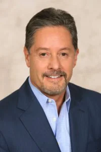 A man in a blue suit and light blue shirt smiling at the camera.