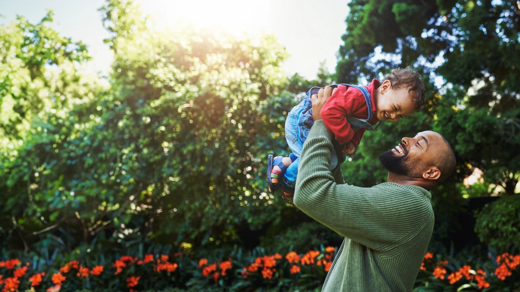 A happy man lifts a smiling toddler in the air in a sunny park with green trees and orange flowers for a webpage about Male Fertility Treatment Options
