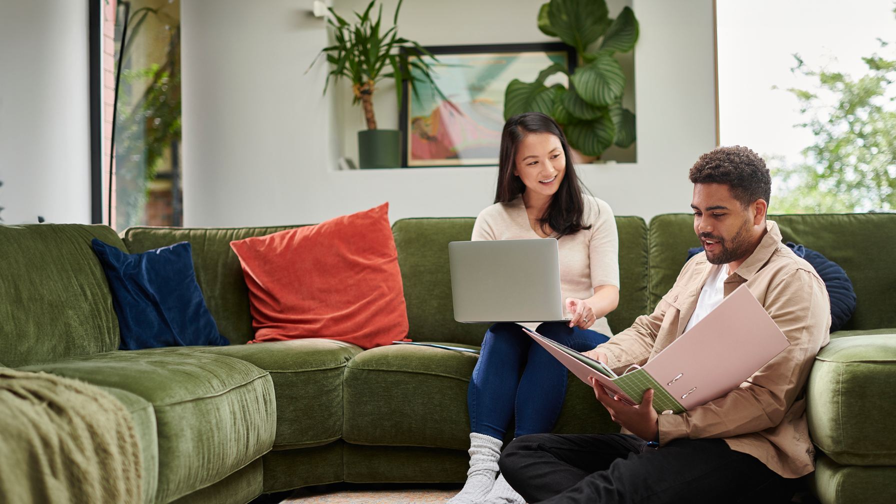 Two people sitting in a cozy living room, working on a laptop and reviewing documents. The room has a green couch with colorful cushions, potted plants, and a window.
