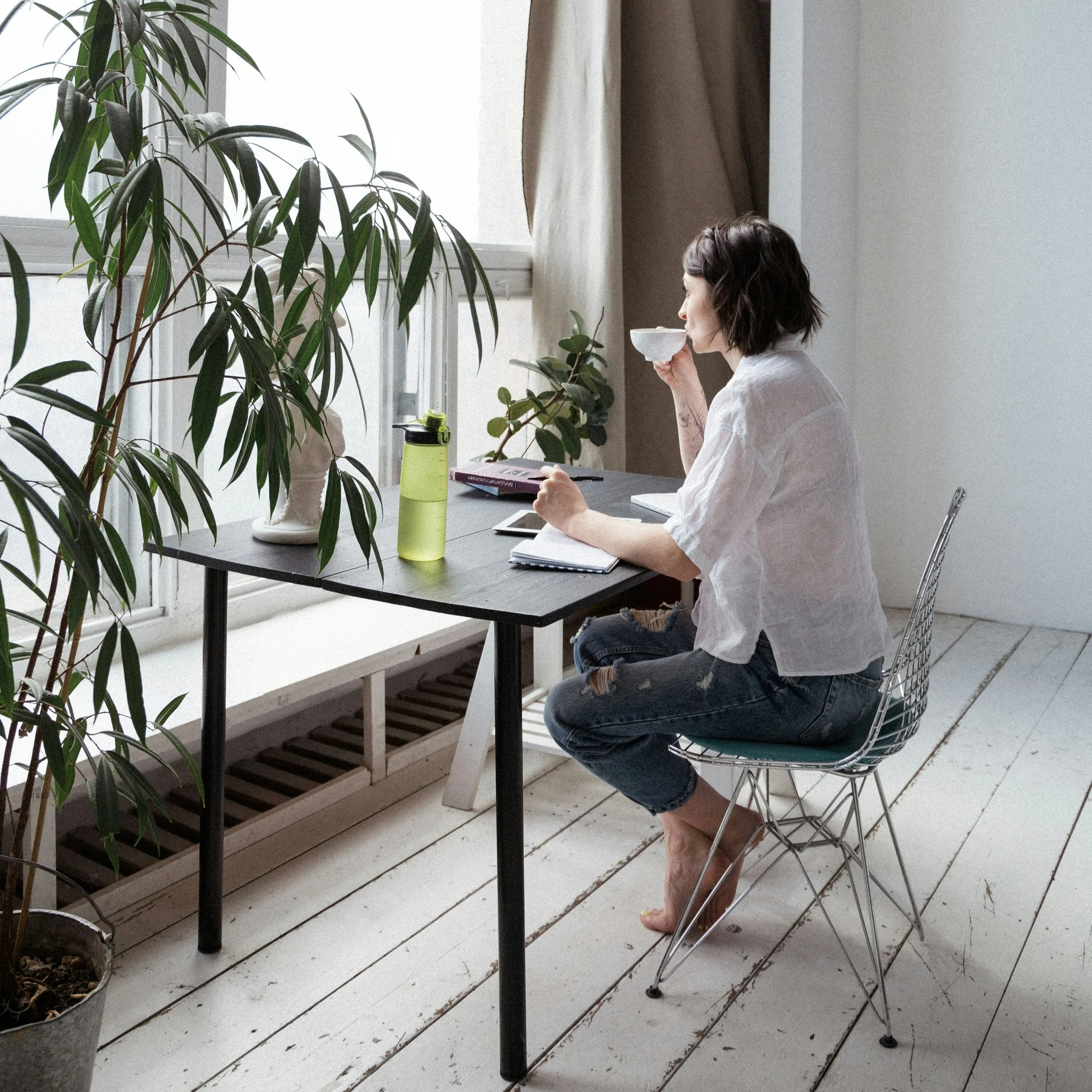 Woman seated at a desk beside a bright window, holding a cup and looking outside