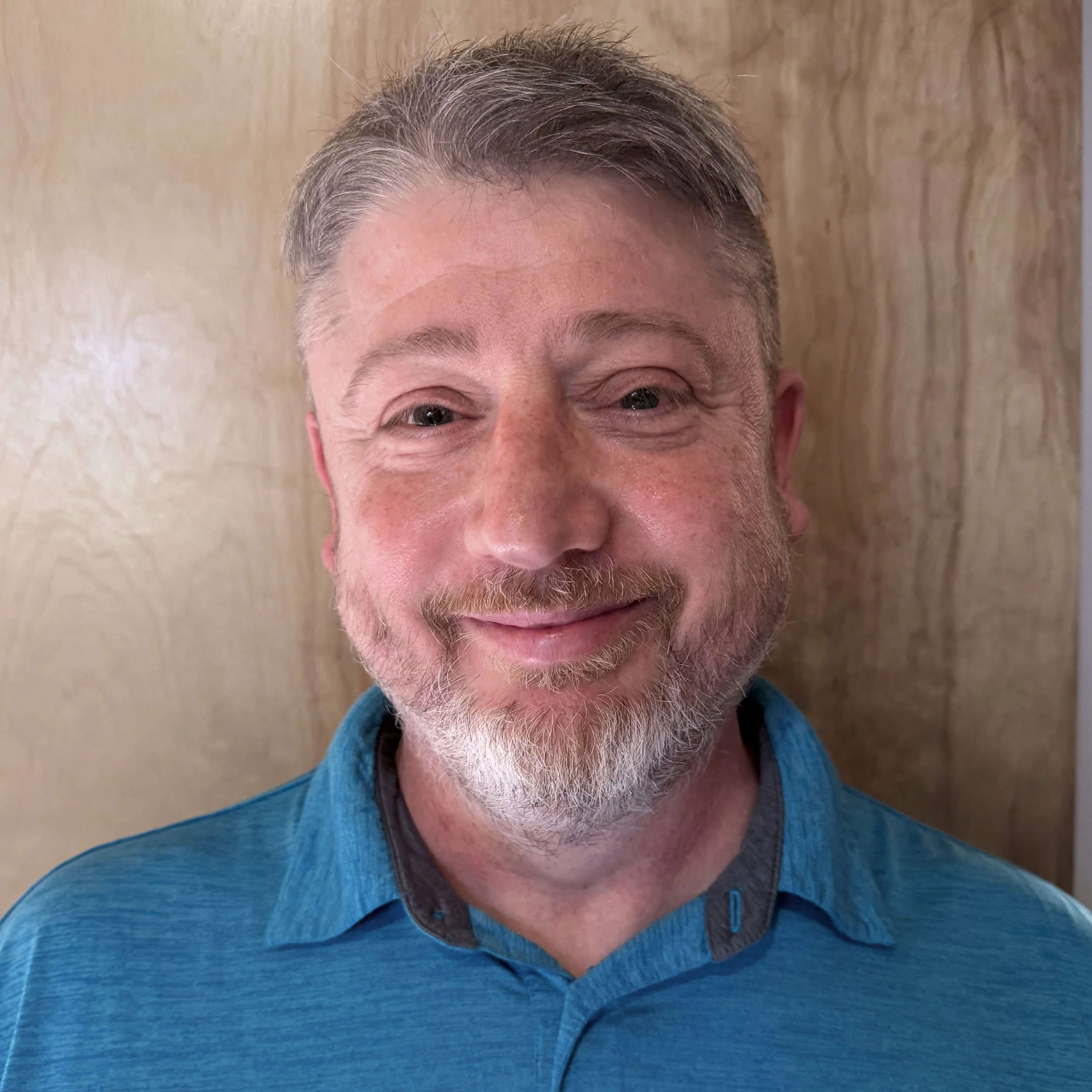 A smiling man with short gray hair and beard, wearing a beige shirt and a patterned tie, standing in front of a Christmas tree.