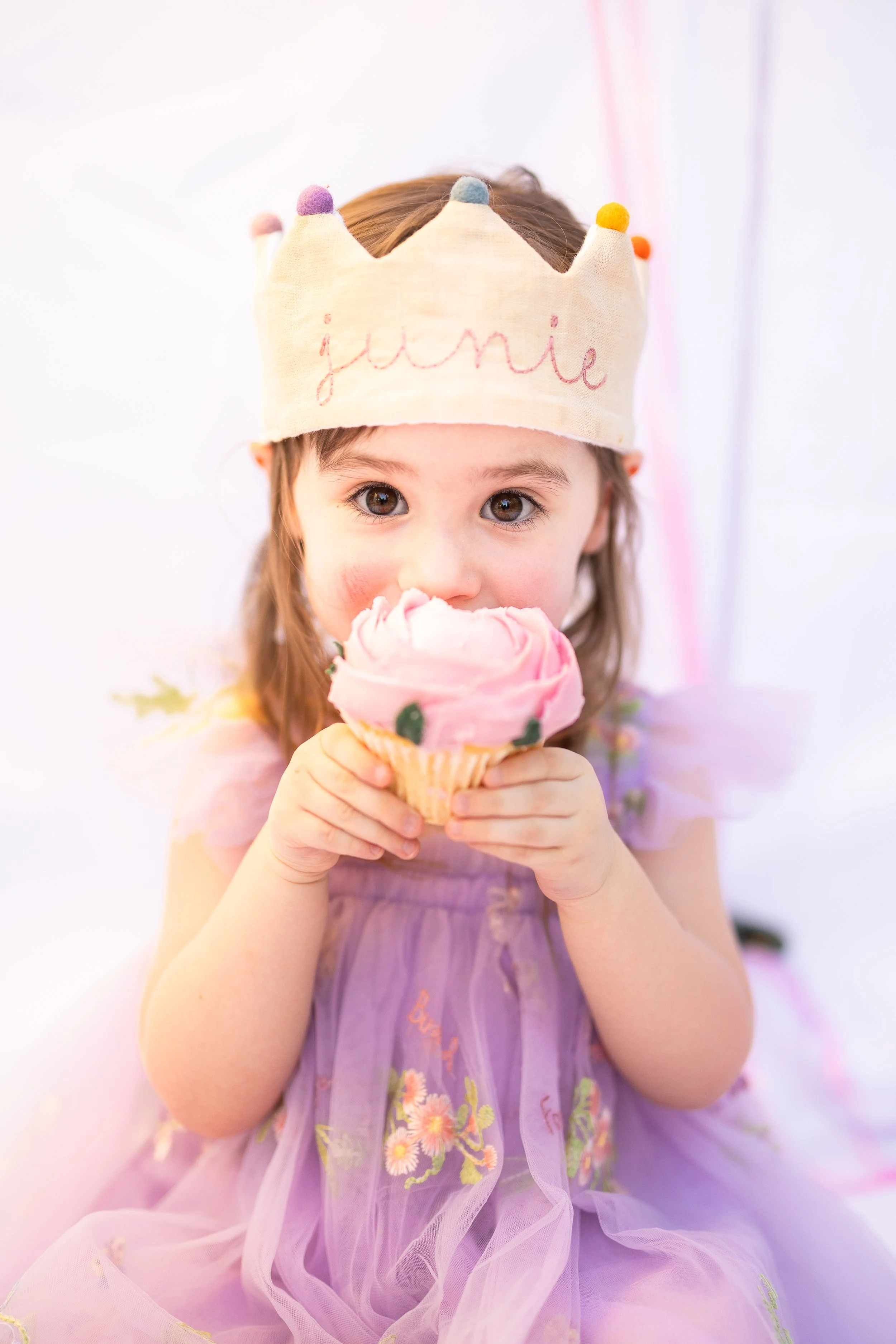 A young girl with brown hair and big eyes, wearing a paper crown with colored pom-poms and the name 'Janie' embroidered on it, is holding and about to eat a pink ice cream cone decorated with small flowers.