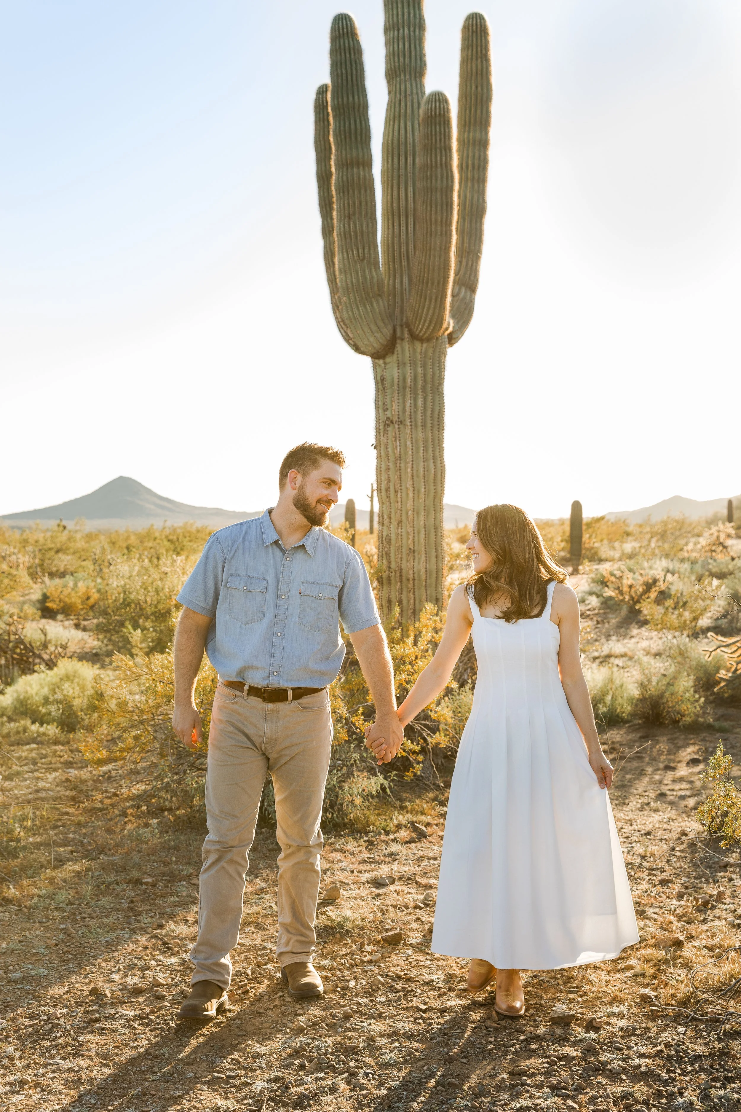 A man and woman holding hands, walking together in a desert landscape with a large saguaro cactus behind them, during sunset.