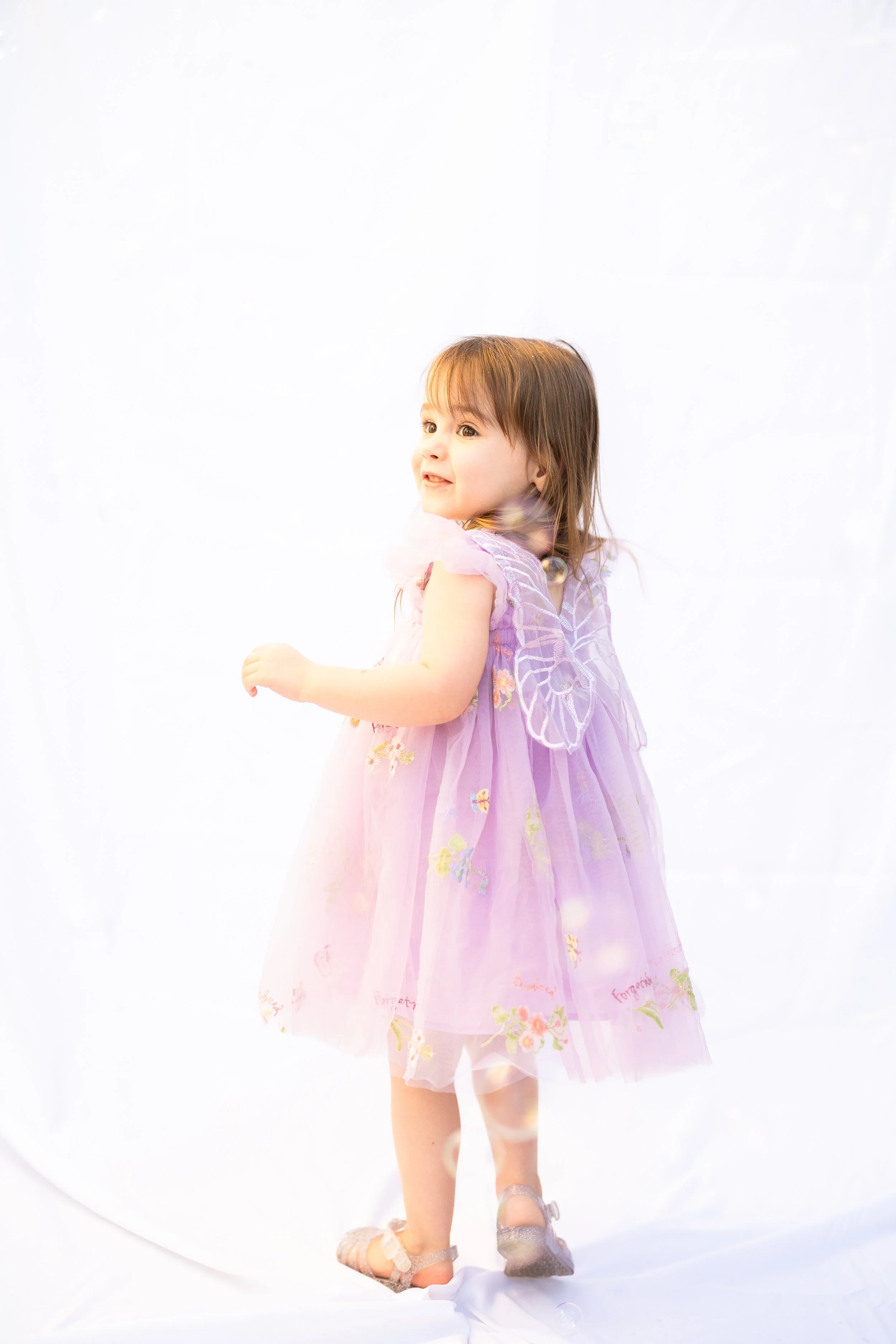 A young girl in a pastel purple dress with floral patterns and sheer overlay, standing against a white background. She is smiling and looking to the side, with light brown hair and wearing sandals.