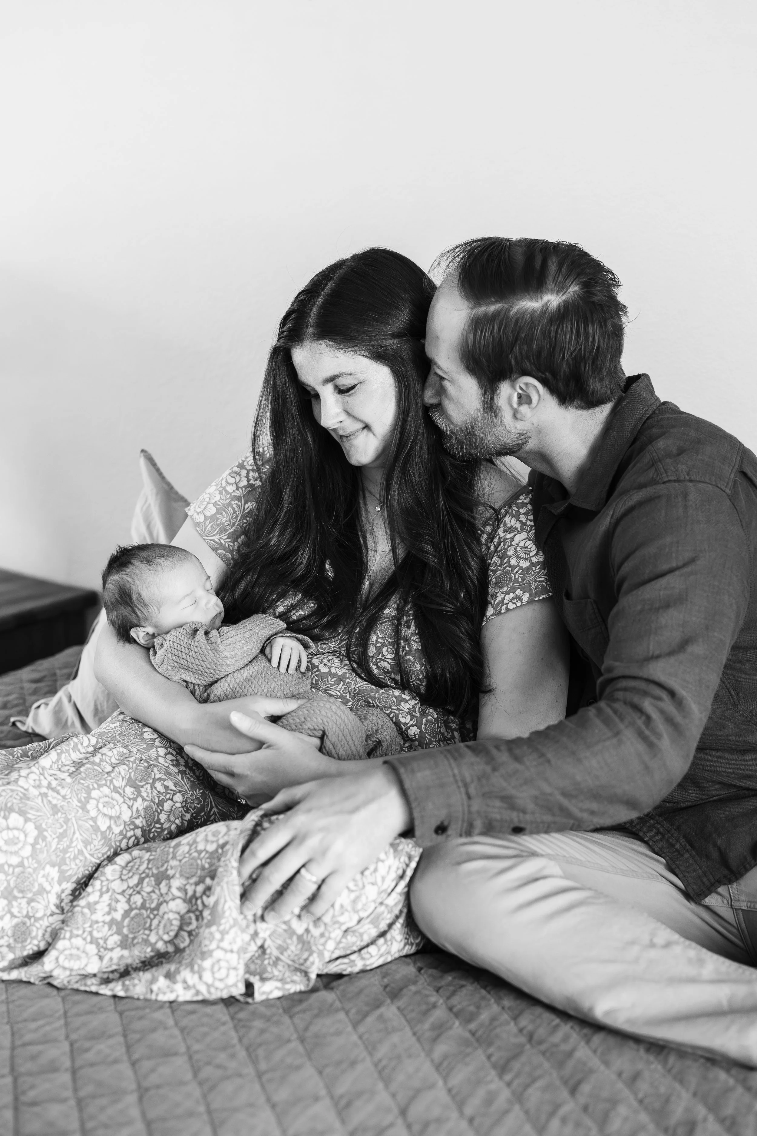 A black and white photo of a family moment with a woman holding a sleeping baby, a man leaning in to kiss her on the forehead, all sitting on a patterned quilt on the floor against a plain wall.
