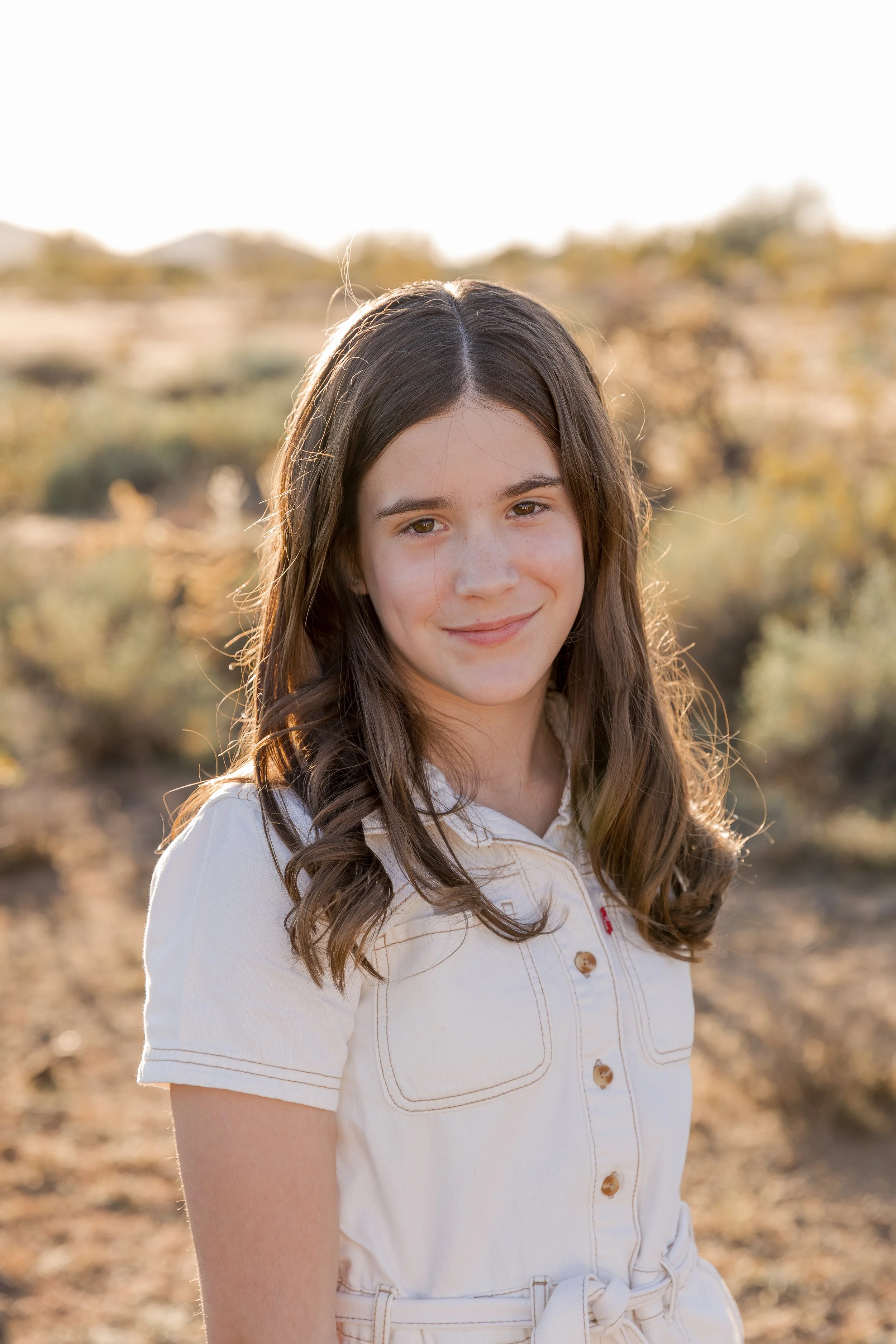 A young girl with long brown hair and light skin smiling outdoors during sunset, wearing a beige short-sleeved dress with buttons and a tied waist, with a desert landscape in the background.