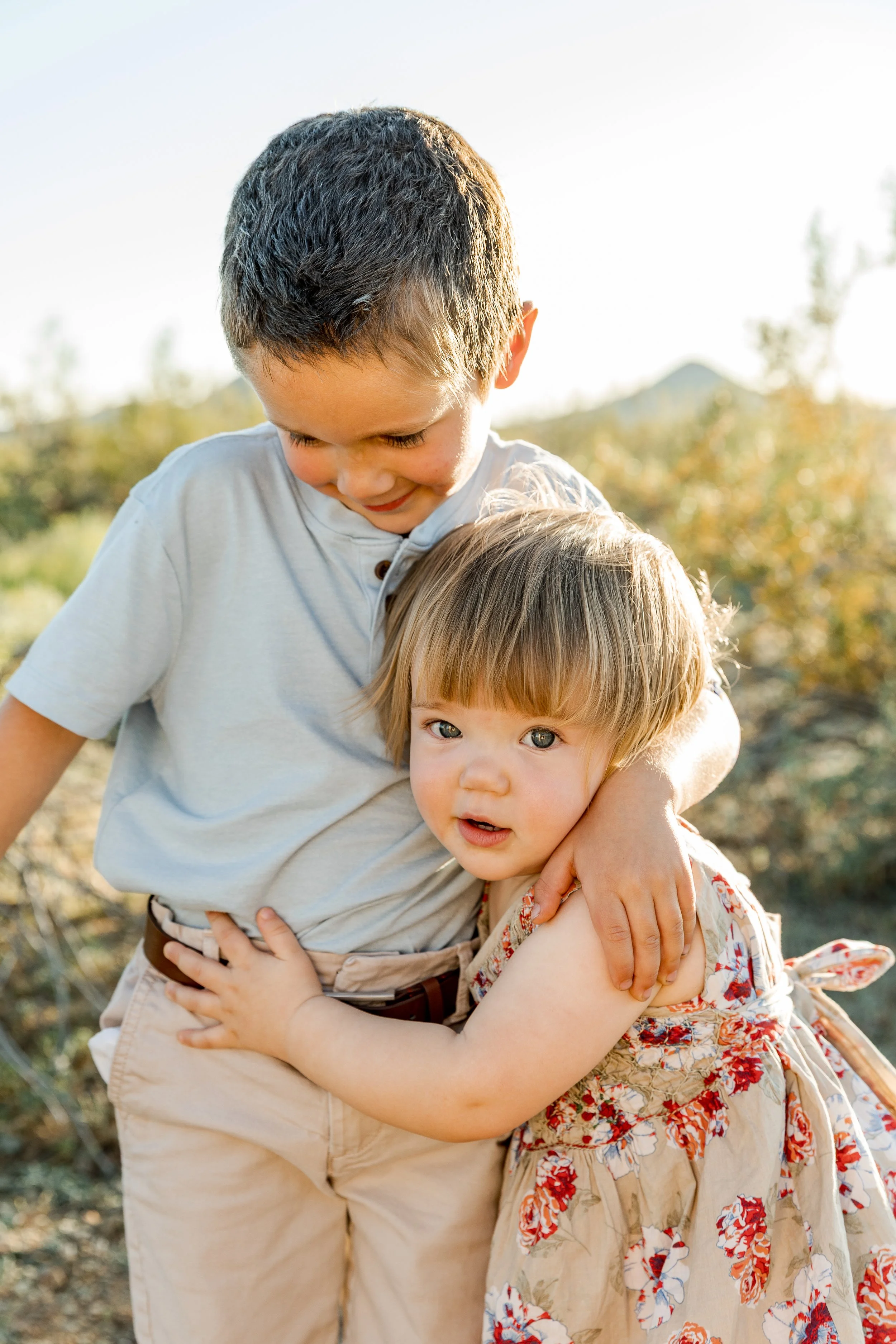 Two young children, a boy and a girl, hugging outdoors on a sunny day with blurred trees and a mountain in the background.