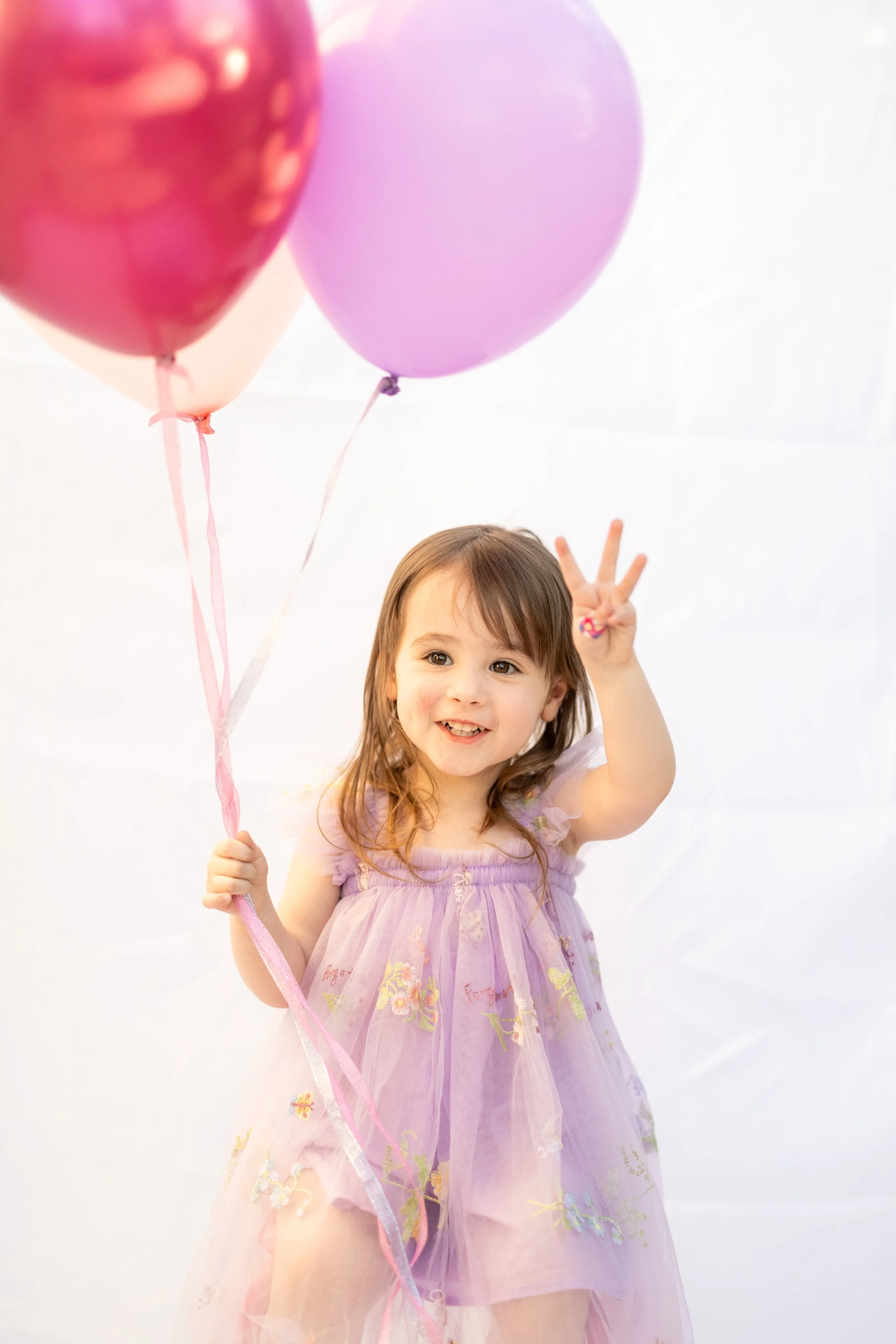 A young girl with brown hair in a purple dress holding pink, purple, and peach balloons and making a peace sign with one hand.