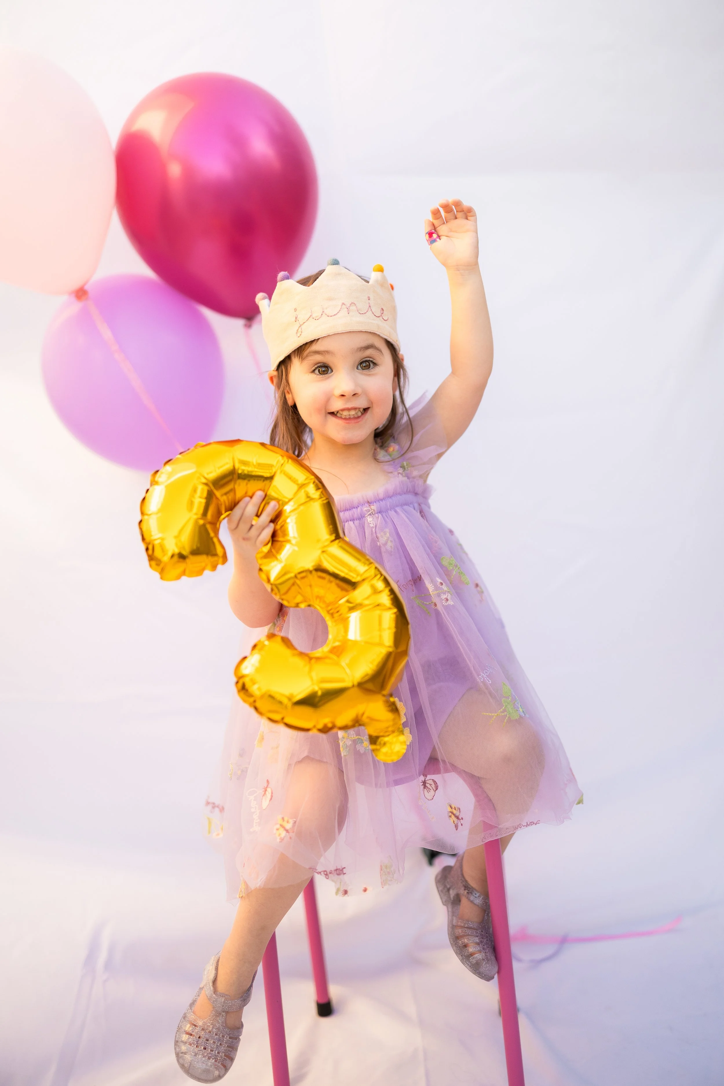 A young girl wearing a crown and a purple dress, holding a gold balloon shaped as the number three, and smiling with her hand raised.