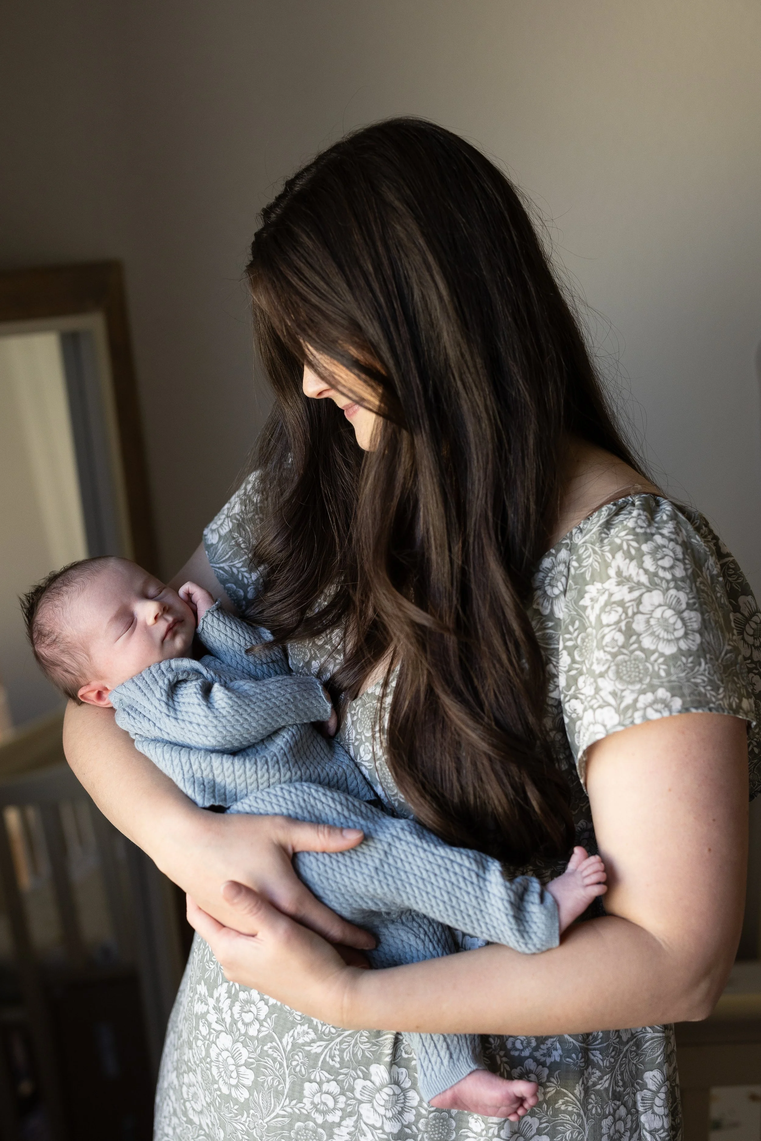 A woman with long, dark brown hair gently holding a sleeping infant. The woman wears a light-colored dress with a floral pattern, and the baby is dressed in a gray textured outfit.