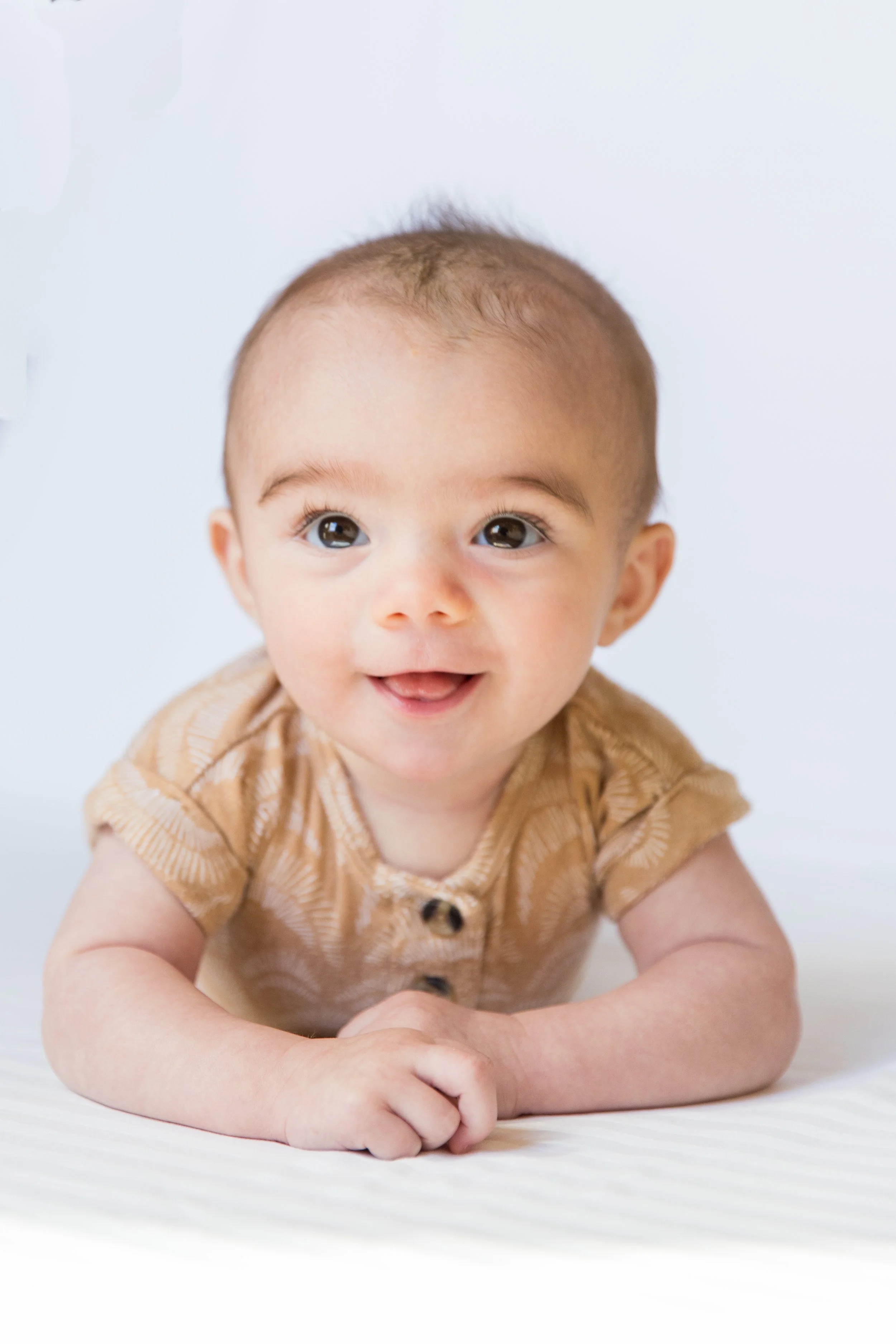 A baby with big brown eyes and light brown hair, wearing a tan and brown patterned shirt, smiling and crawling on a white surface against a white background.