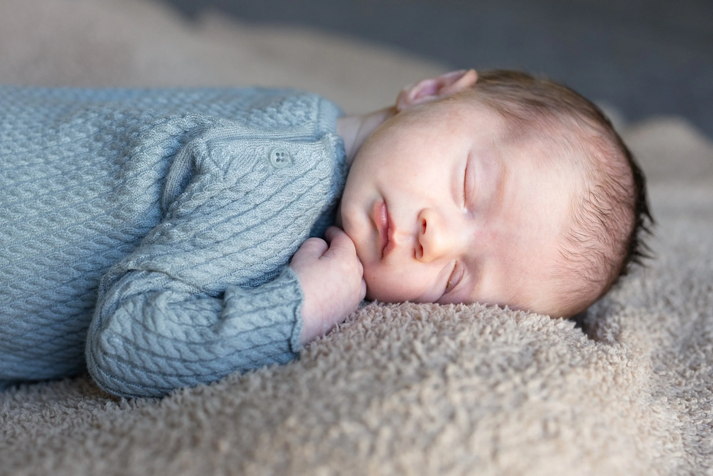 A sleeping baby lying on a soft beige carpet, dressed in a textured blue sweater, with eyes closed and hand near the mouth.