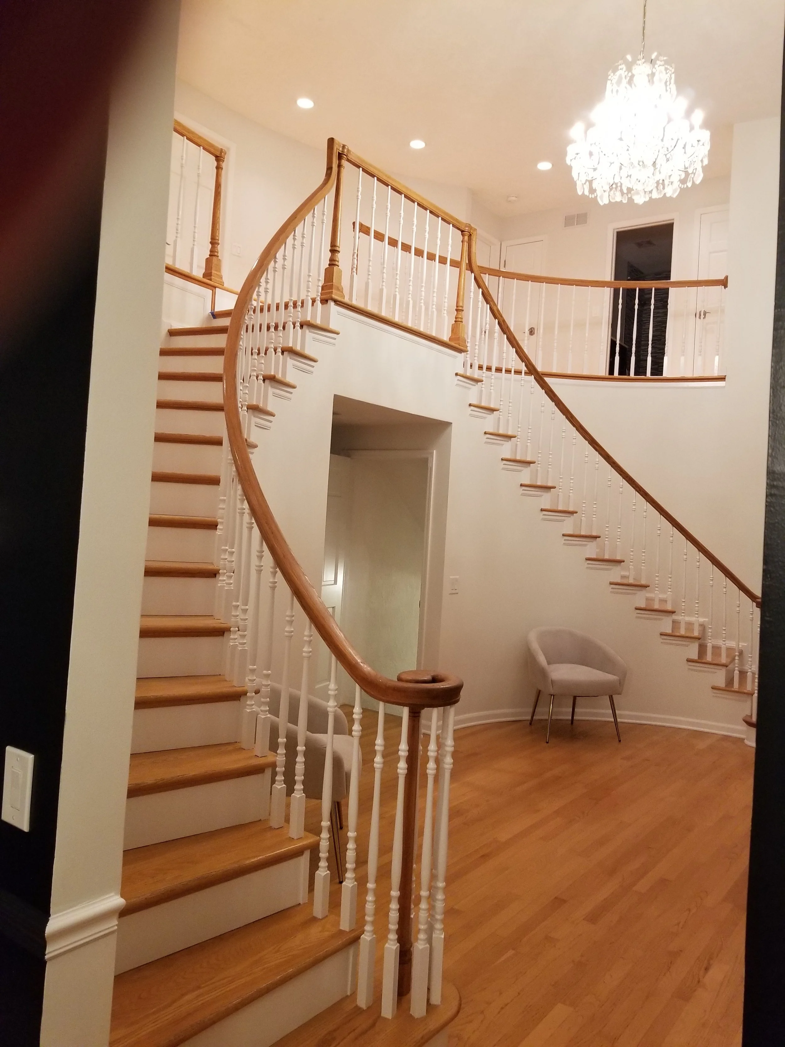 A distant hallway viewpoint looking into an expansive entryway. The graceful curve of the grand staircase is accentuated by the clean white painted spindles and risers, contrasting the natural wood treads and railing. The black accent wall of the adj