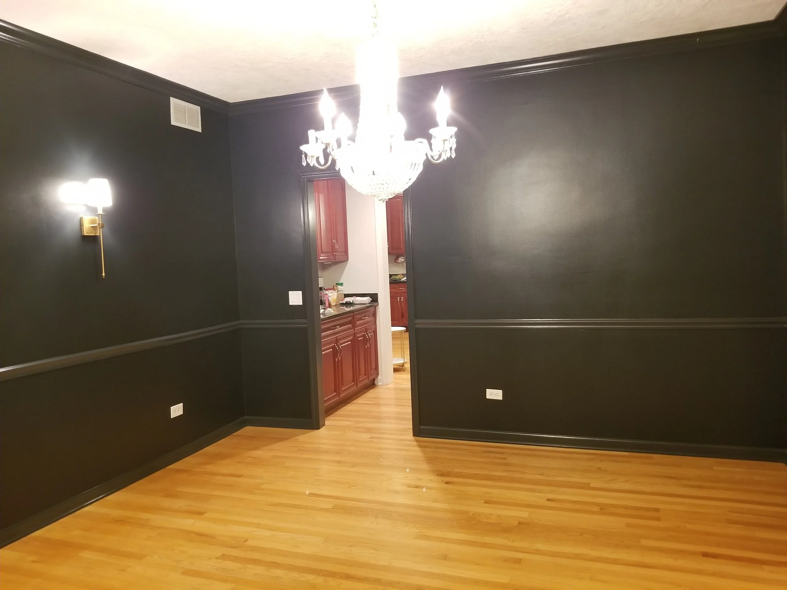 A dramatic and modern transformation of a dining area featuring deep matte black walls. Bright white crystal chandeliers and wall sconces pop against the dark paint, highlighting clean lines and elegant contrasting wainscoting. Through the open doorw