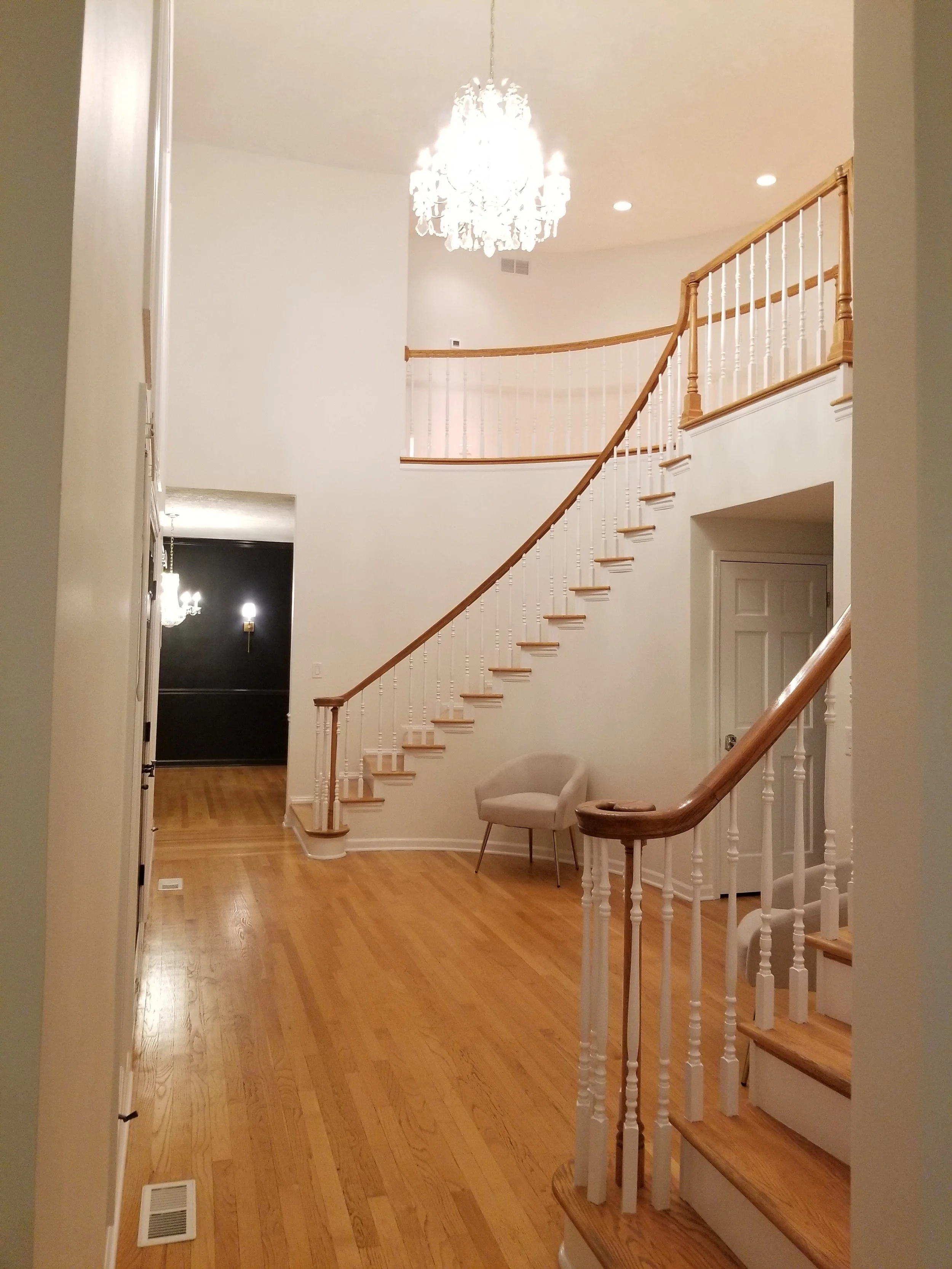 A spectacular, wide-angle view of a luxurious two-story foyer. The elegant curved grand staircase, completely finished with professional paint and wood stain, leads the eye up towards a sparkling crystal chandelier and recess lighting. A modern velve