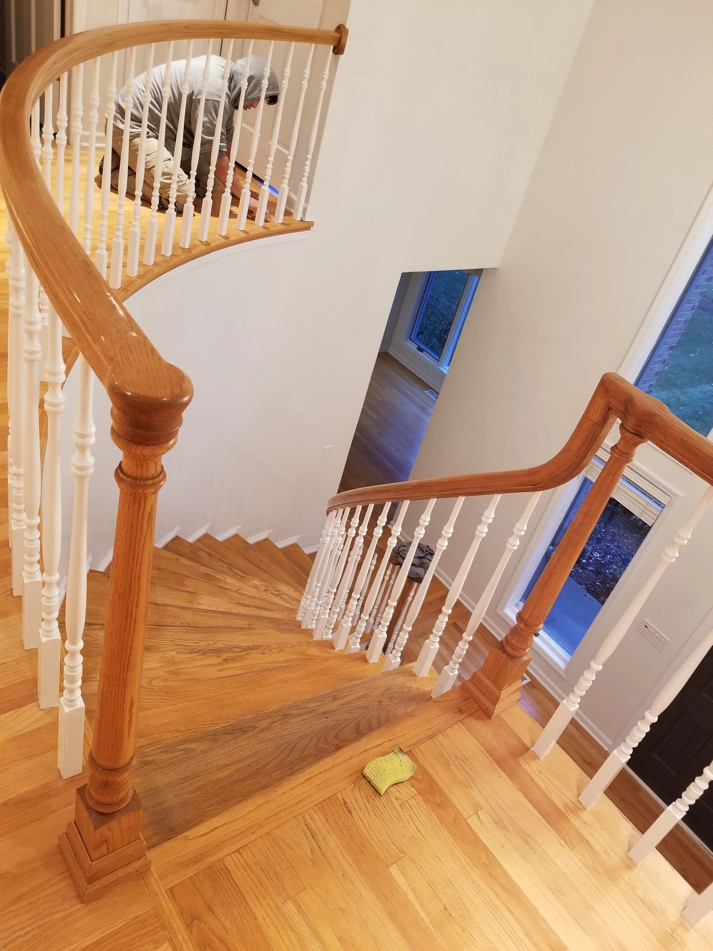 Close-up view looking down a grand, curved staircase. A skilled worker in a hoodie meticulously applies a fresh coat of white paint to the intricate balusters. The rich, light wood steps and handrail show high-gloss finish. A sanding sponge rests on 