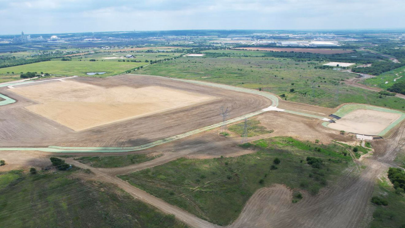 Aerial view of a large construction site with cleared land, dirt roads, and green areas, with a city skyline in the distance under a partly cloudy sky.