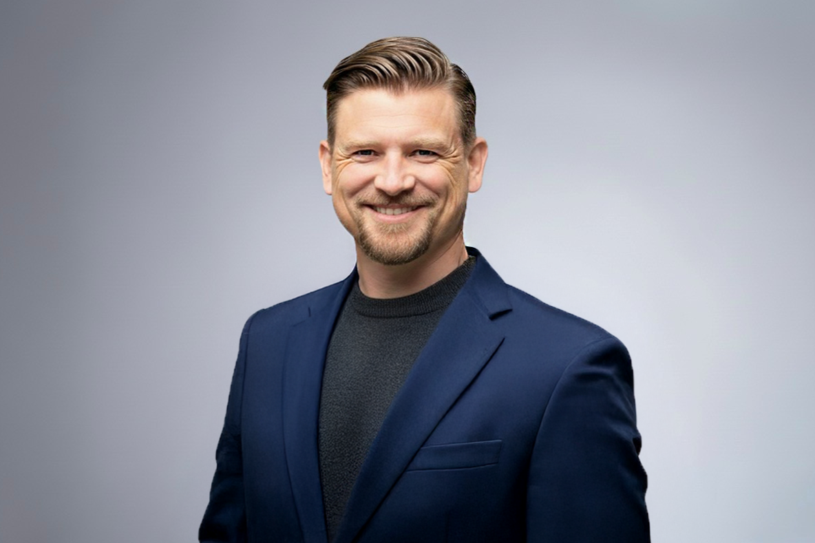 Man with short hair, beard, and mustache wearing a dark blue suit jacket over a black shirt, smiling and standing against a plain light gray background.