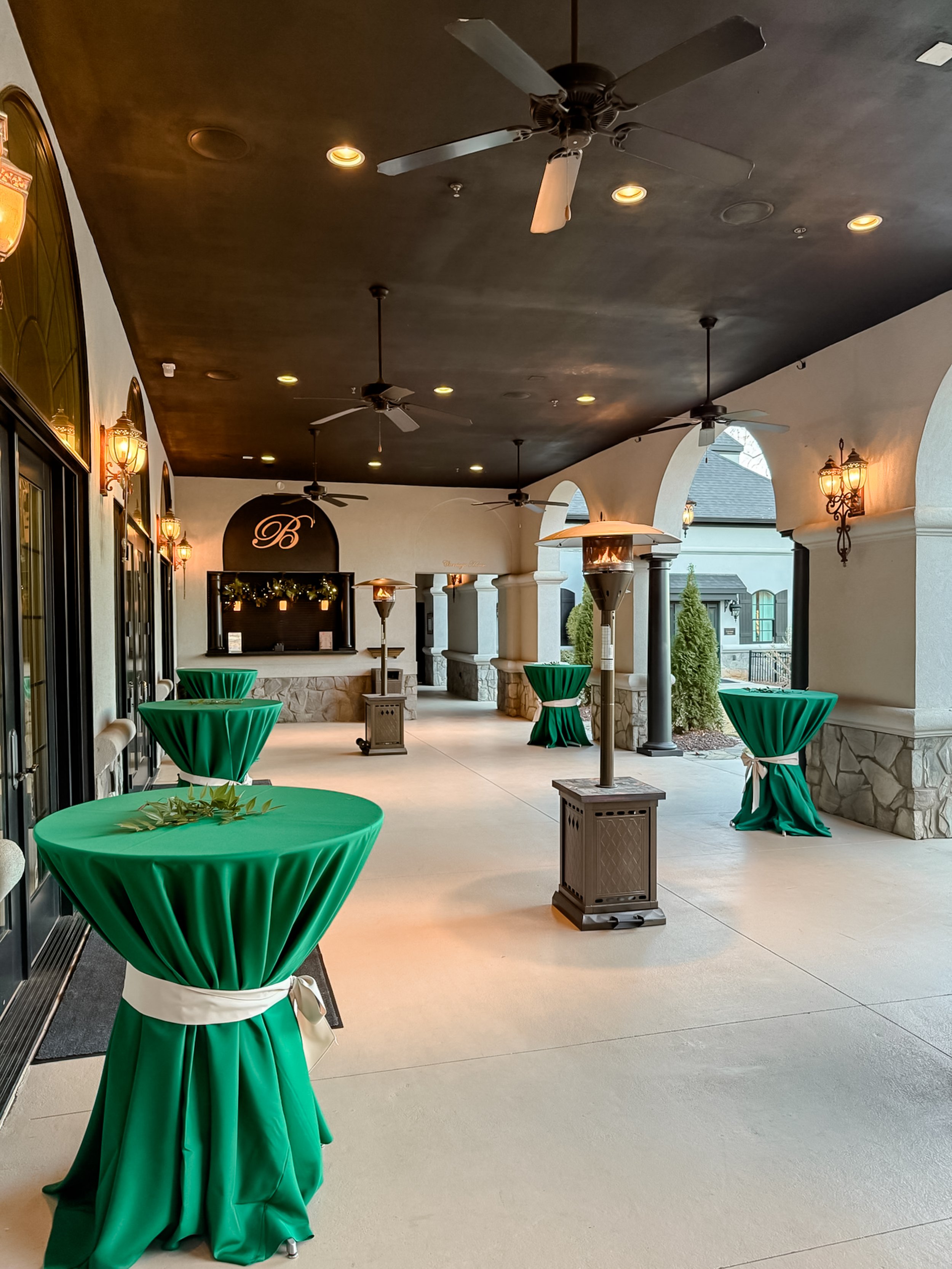 Outdoor covered patio with high-top tables draped in green tablecloths with white sashes, ceiling fans, wall-mounted lantern lights, and a heater, set up for a social event.