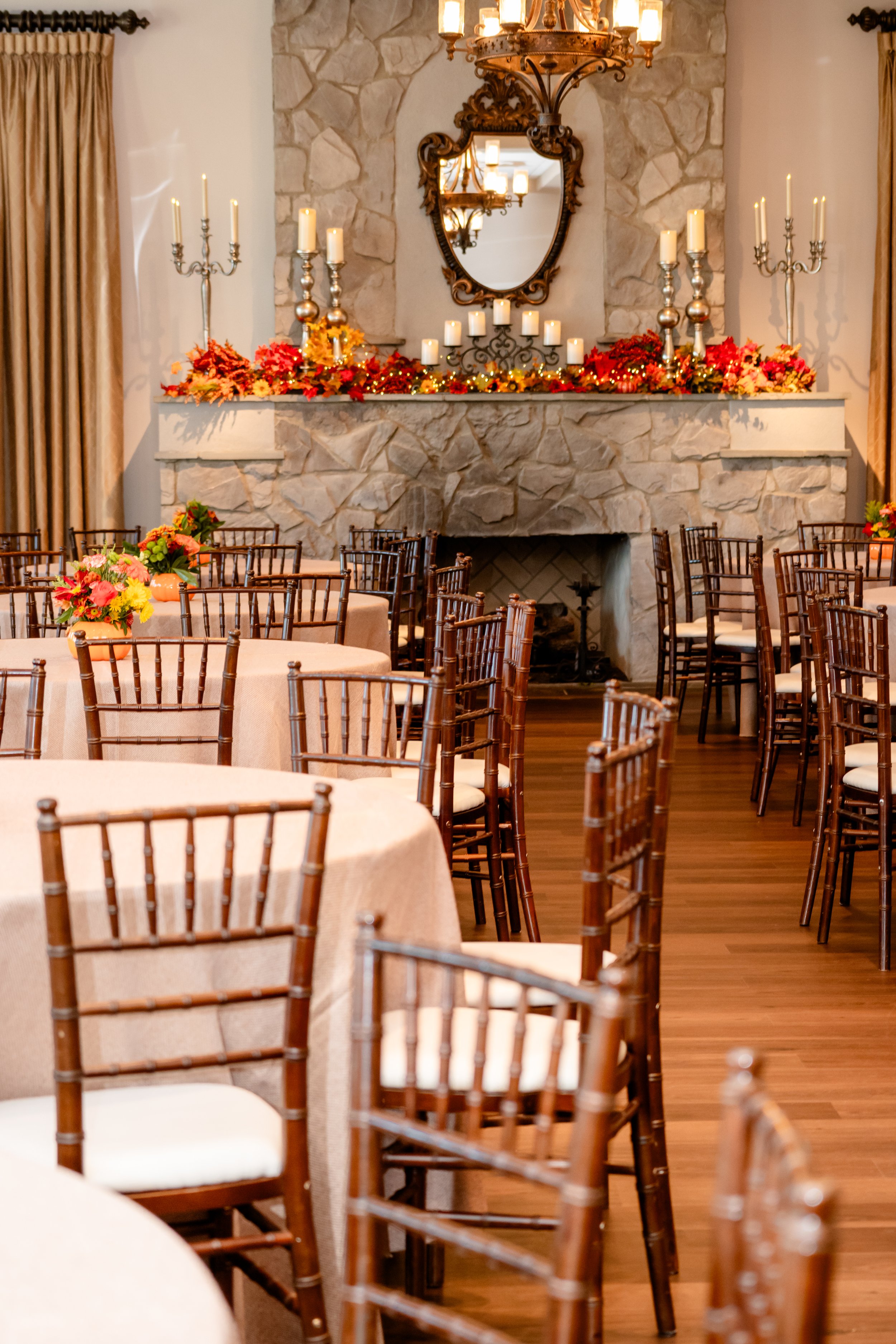 Interior of an elegant event space with round tables covered in beige tablecloths, decorated with colorful flower centerpieces, and surrounded by brown wooden chairs. There is a stone fireplace decorated with candles, flowers, and autumn-themed folia