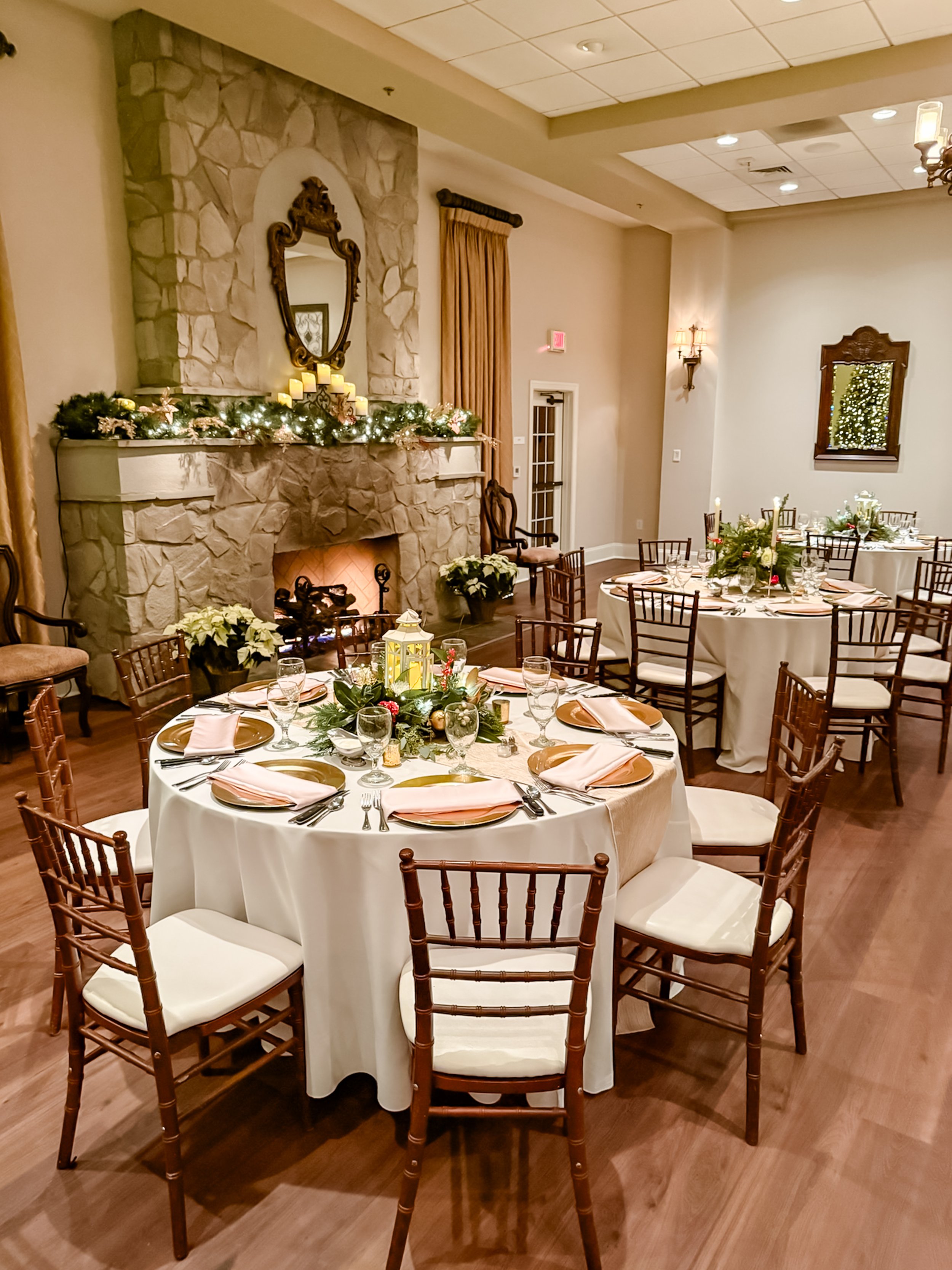 Elegant holiday reception room with round tables, white tablecloths, and festive centerpieces, a stone fireplace decorated with candles and greenery, and warm lighting.