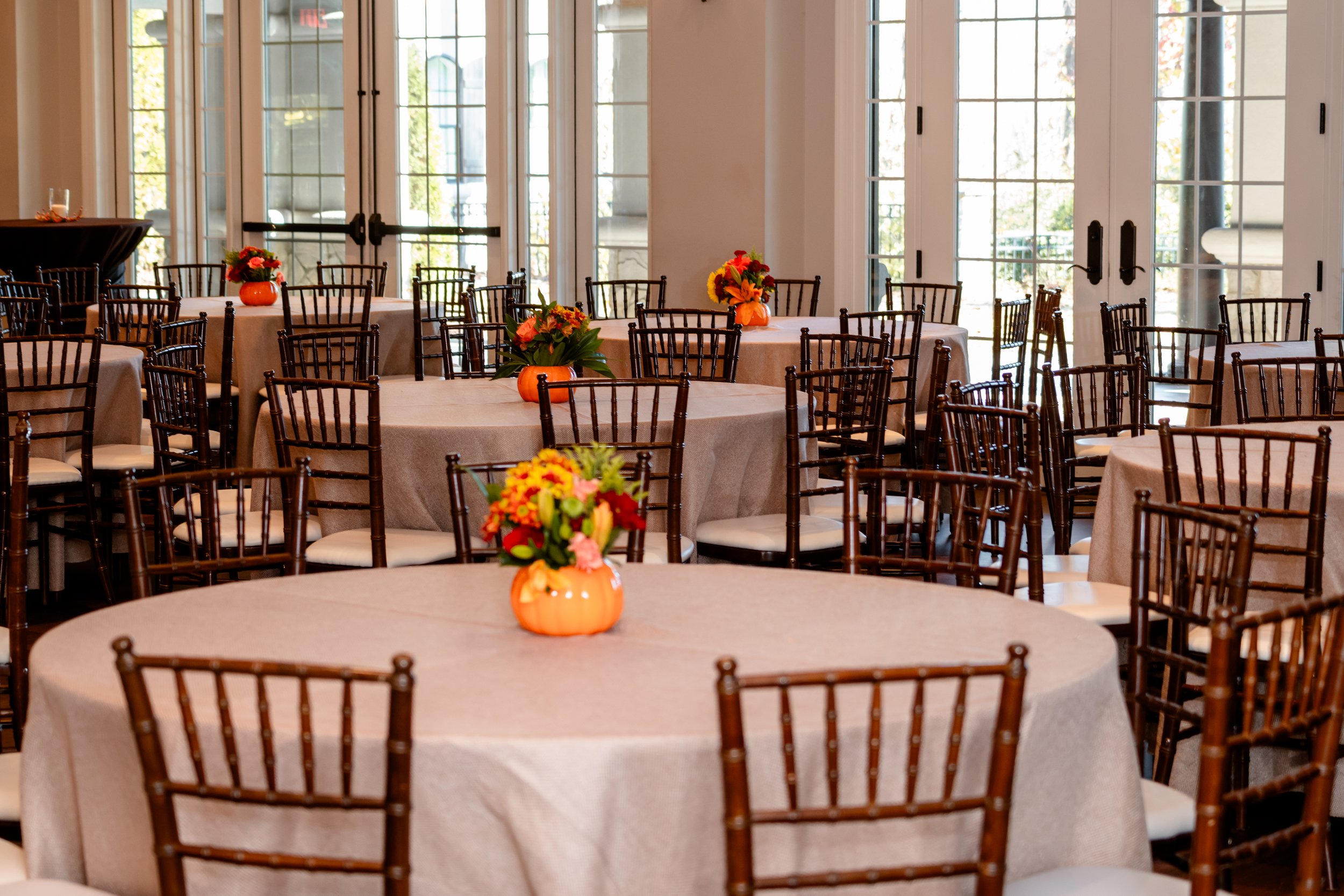 Restaurant or event space with round tables covered in beige tablecloths, decorated with orange pumpkin-shaped vases filled with colorful flowers, surrounded by dark wooden chairs with white cushions, and large glass doors in the background letting i