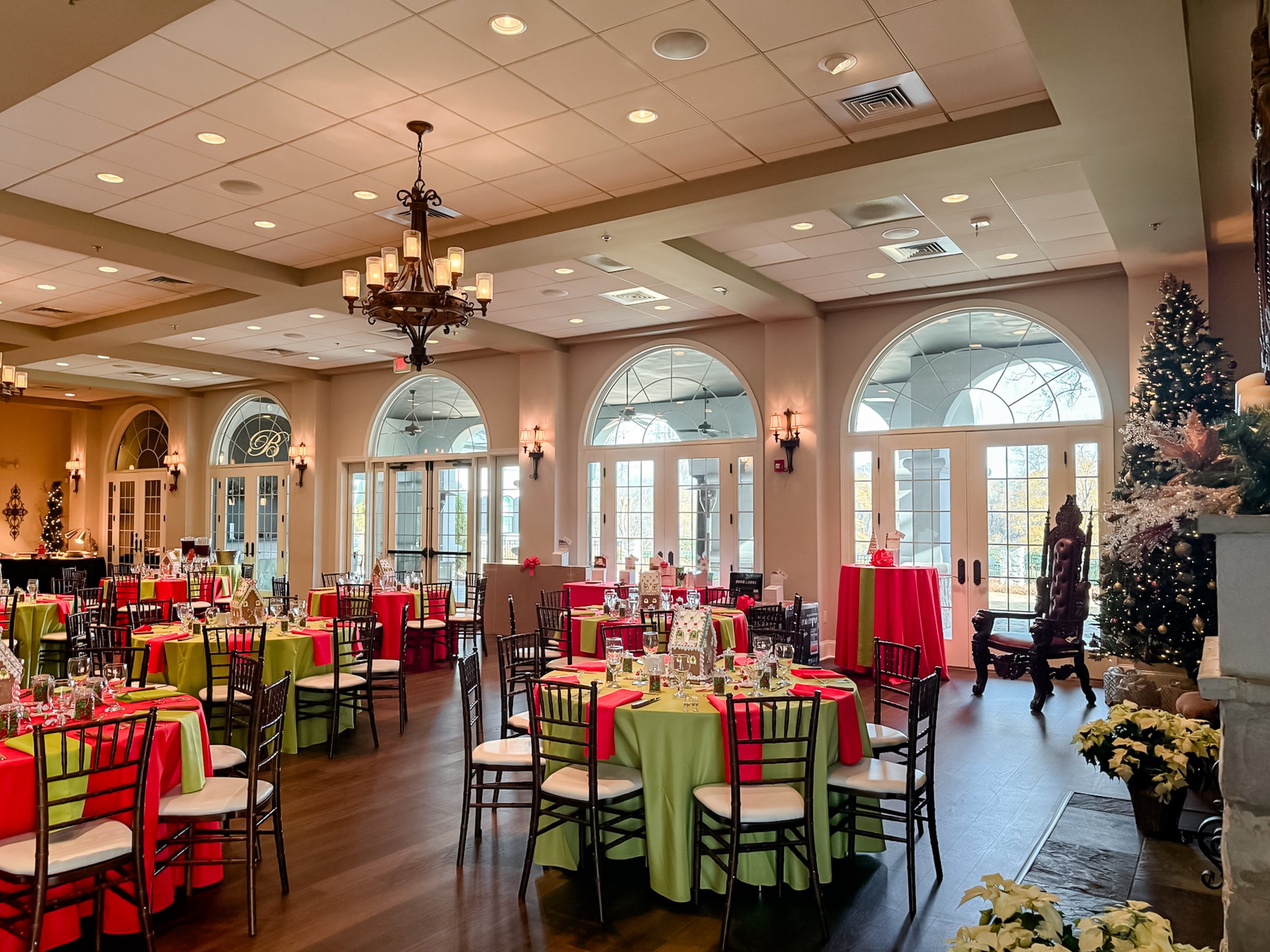 A decorated banquet hall with round tables covered in red and green tablecloths, set with glassware and centerpieces, and decorated for Christmas with a Christmas tree and poinsettias, featuring large arched windows and a chandelier.