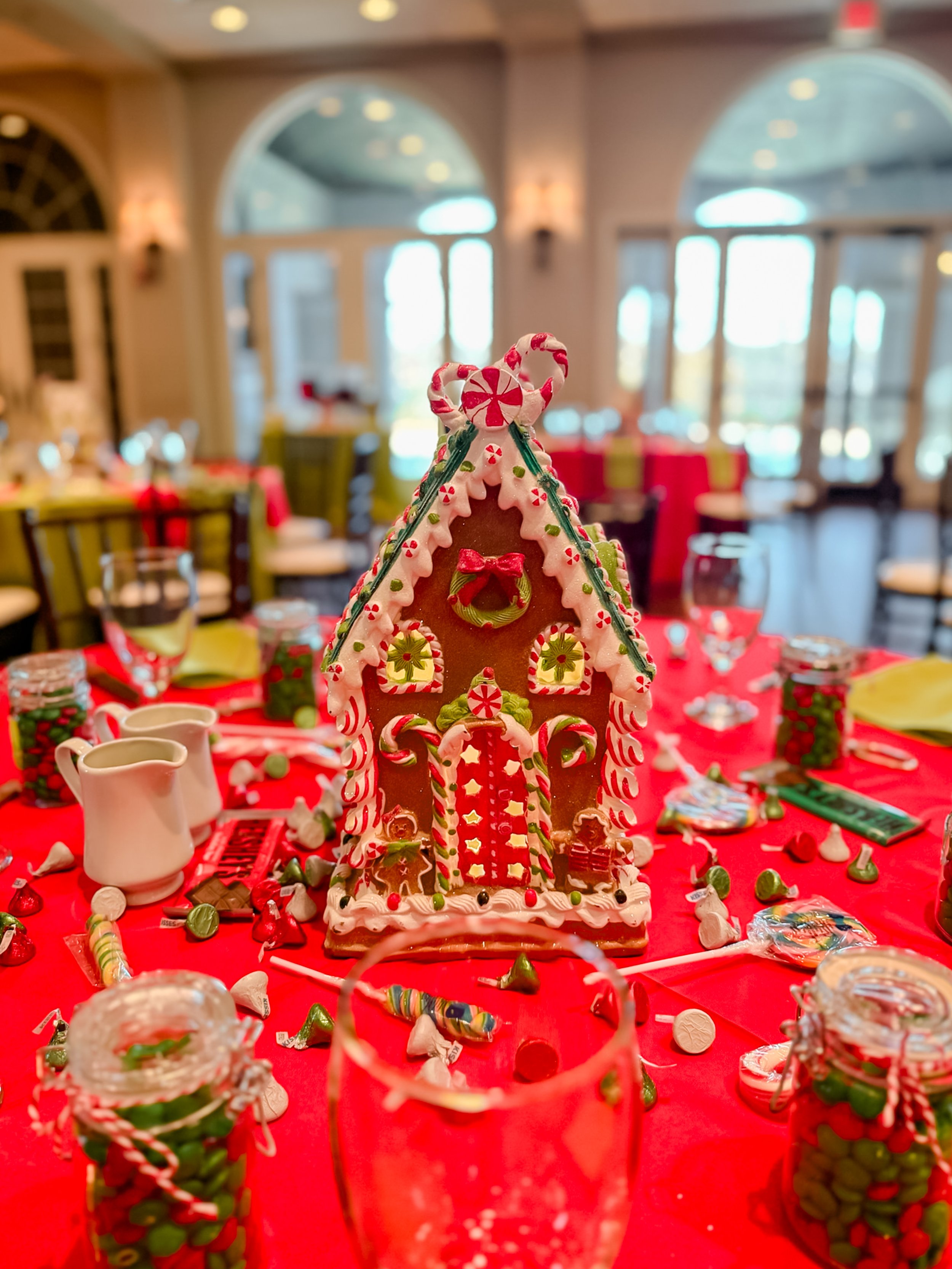 Decorated gingerbread house centerpiece on a red tablecloth in a festive, well-lit dining room with holiday decorations.