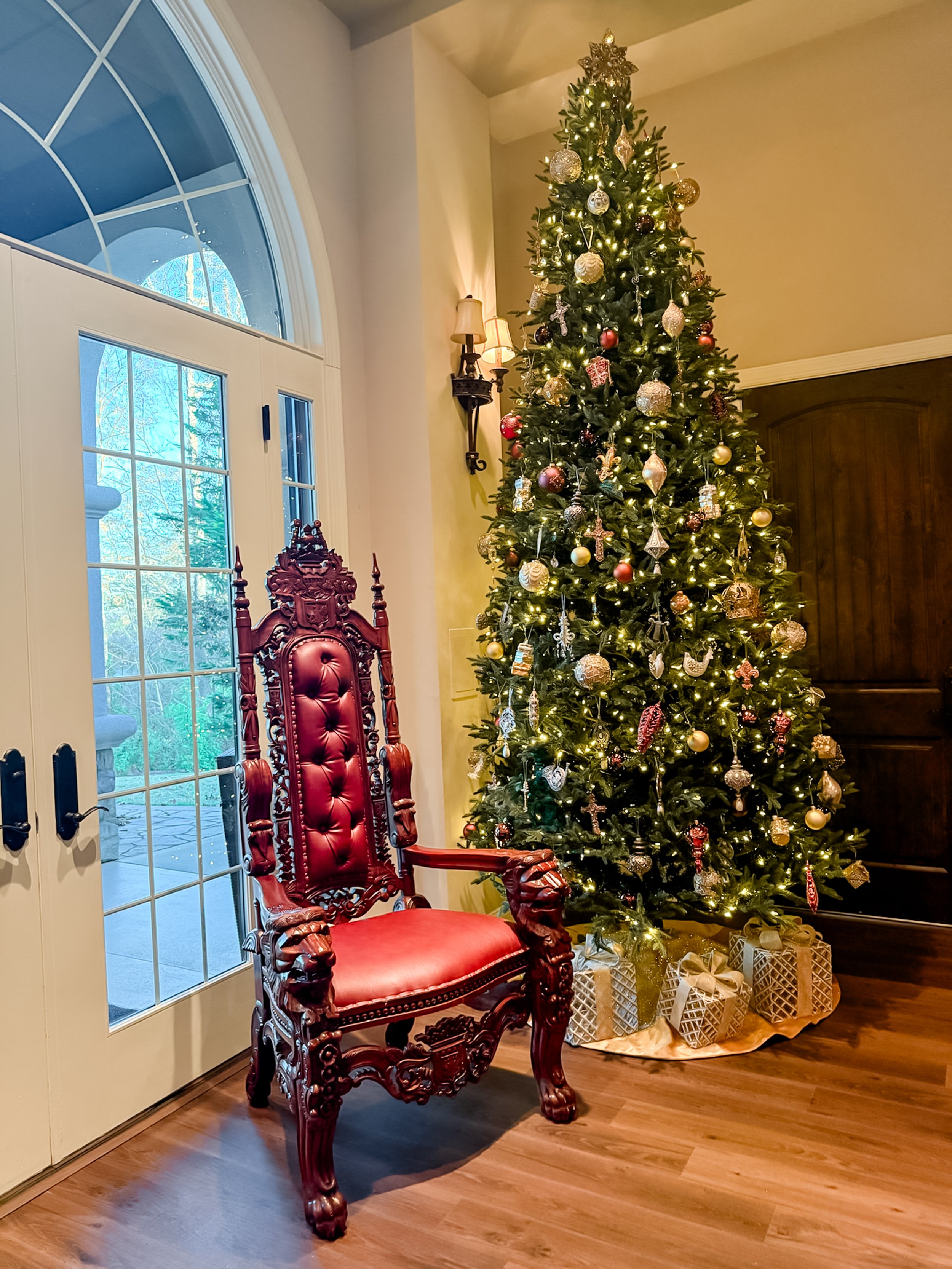 Decorated Christmas tree with ornaments and gifts beneath, beside a carved wooden throne-like chair near glass doors.