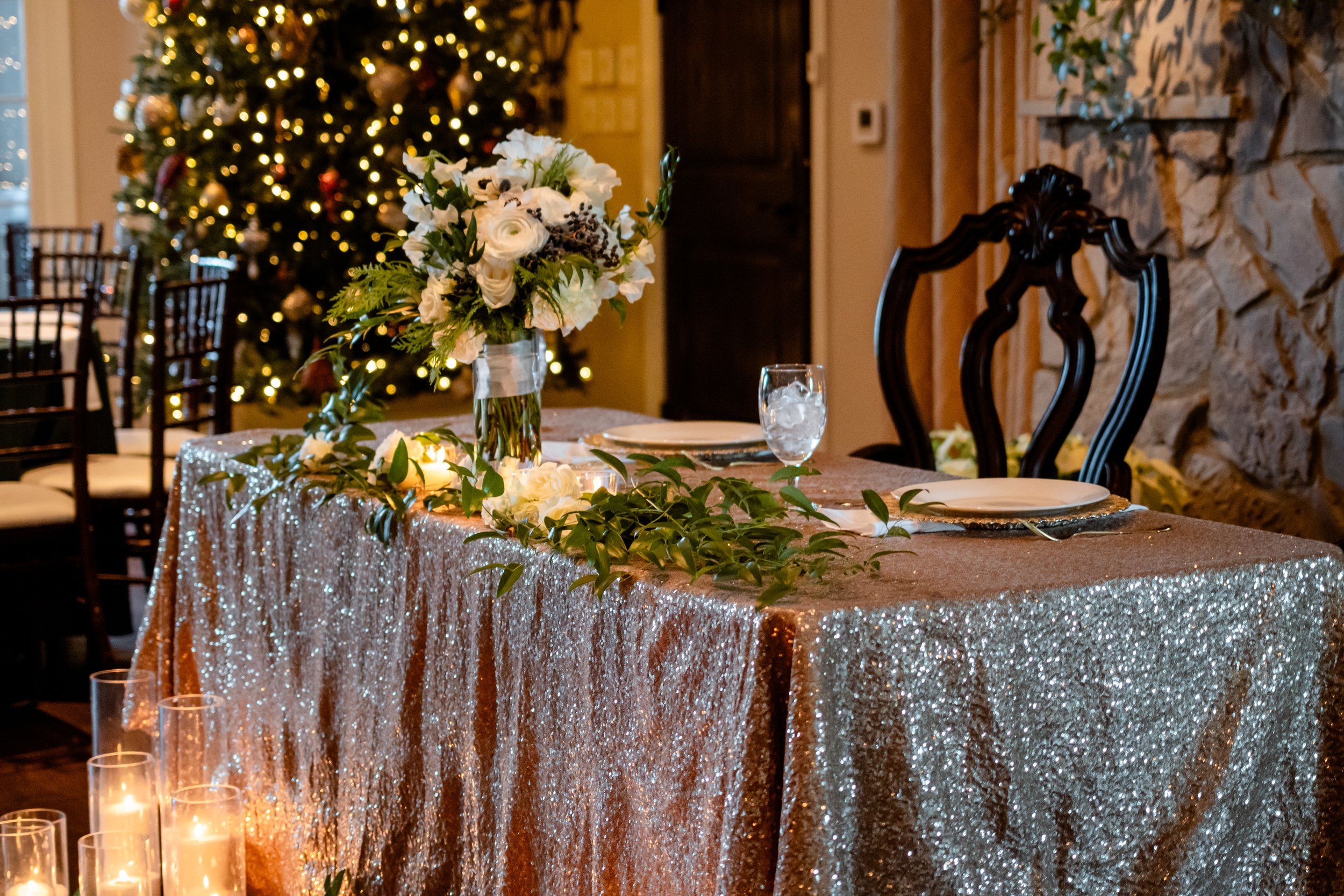 Decorated dining table with a glittery gold tablecloth, a floral centerpiece, and lit candles in a room with a Christmas tree in the background.
