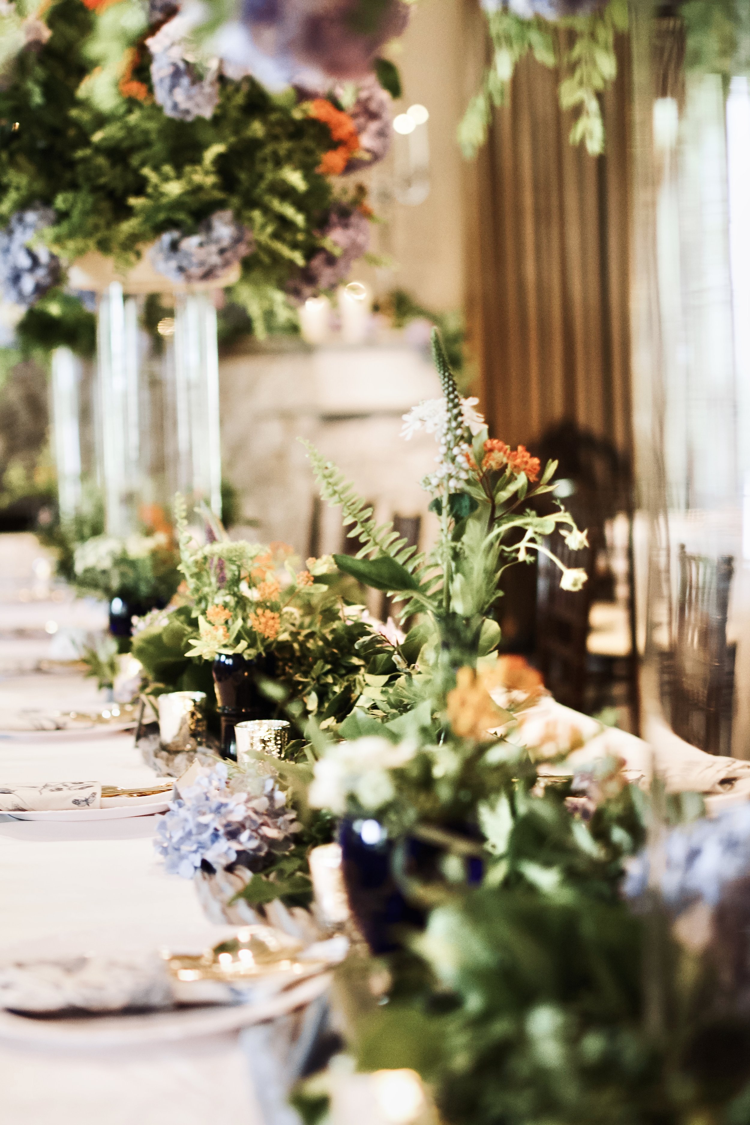 A close-up of a decorated dining table with floral centerpieces, candles, and tableware in a warmly lit room with draped curtains.