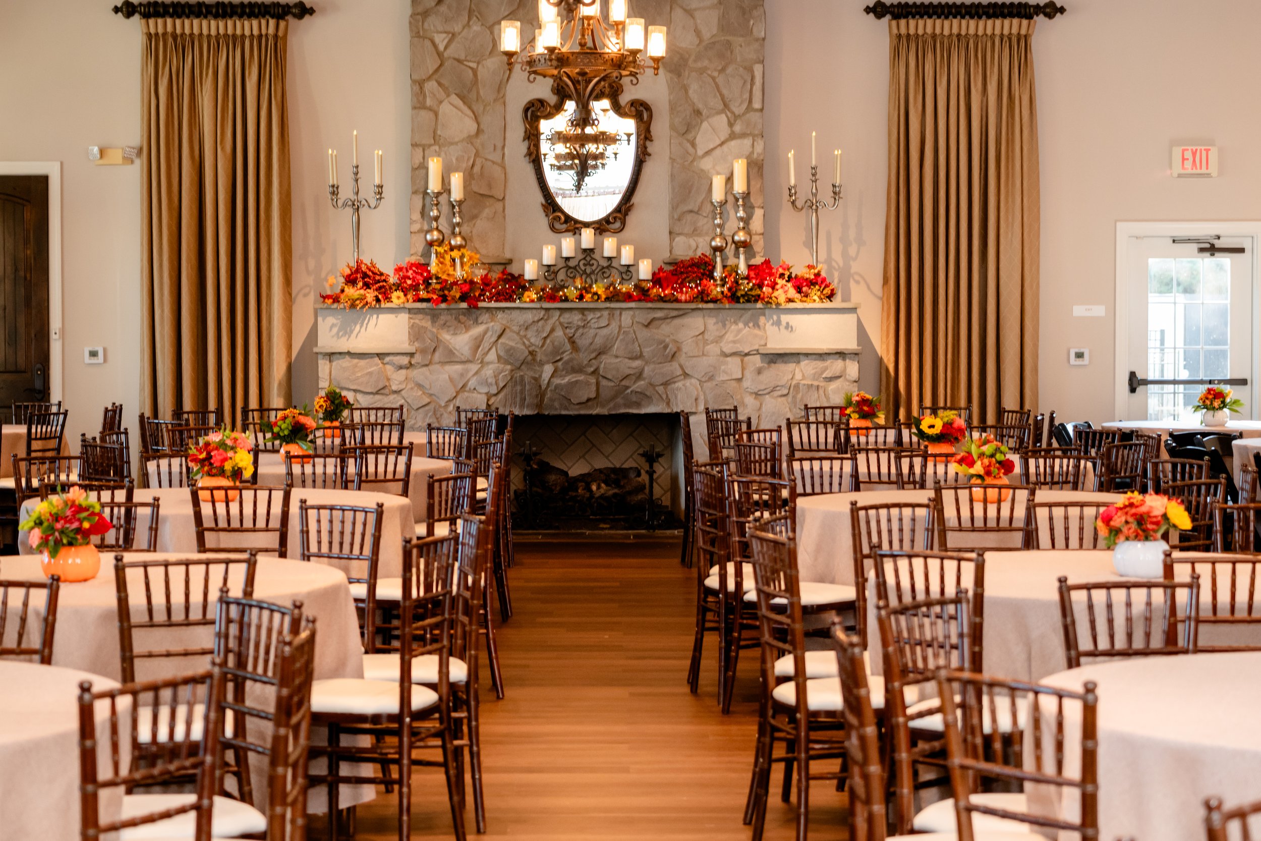 An elegantly decorated banquet hall with round tables covered in beige tablecloths, each adorned with small flower arrangements, and a stone fireplace decorated with candles and fall foliage in the background.
