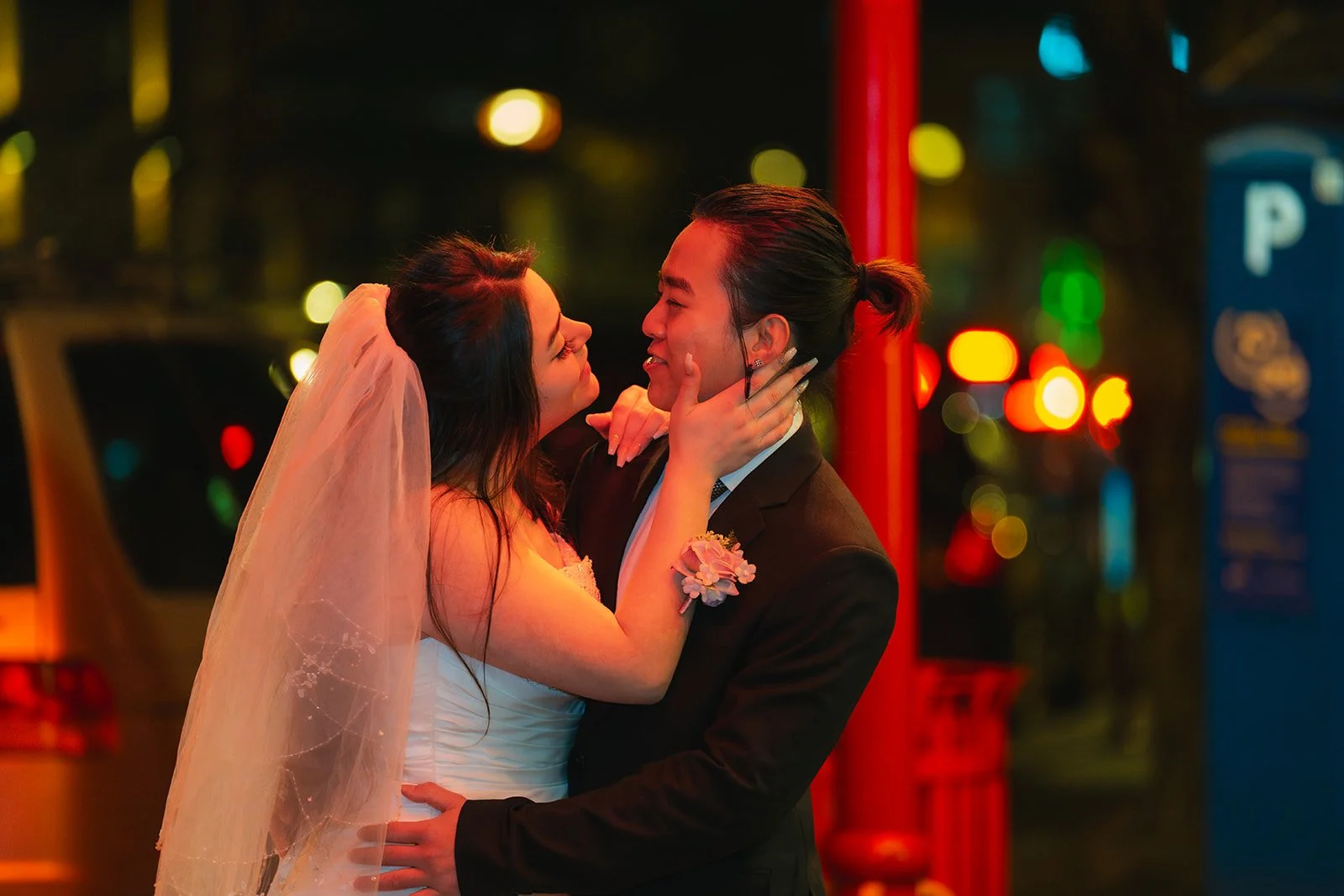 A bride and groom wearing wedding attire sharing a romantic moment on a city street at night with blurred colorful city lights in the background.