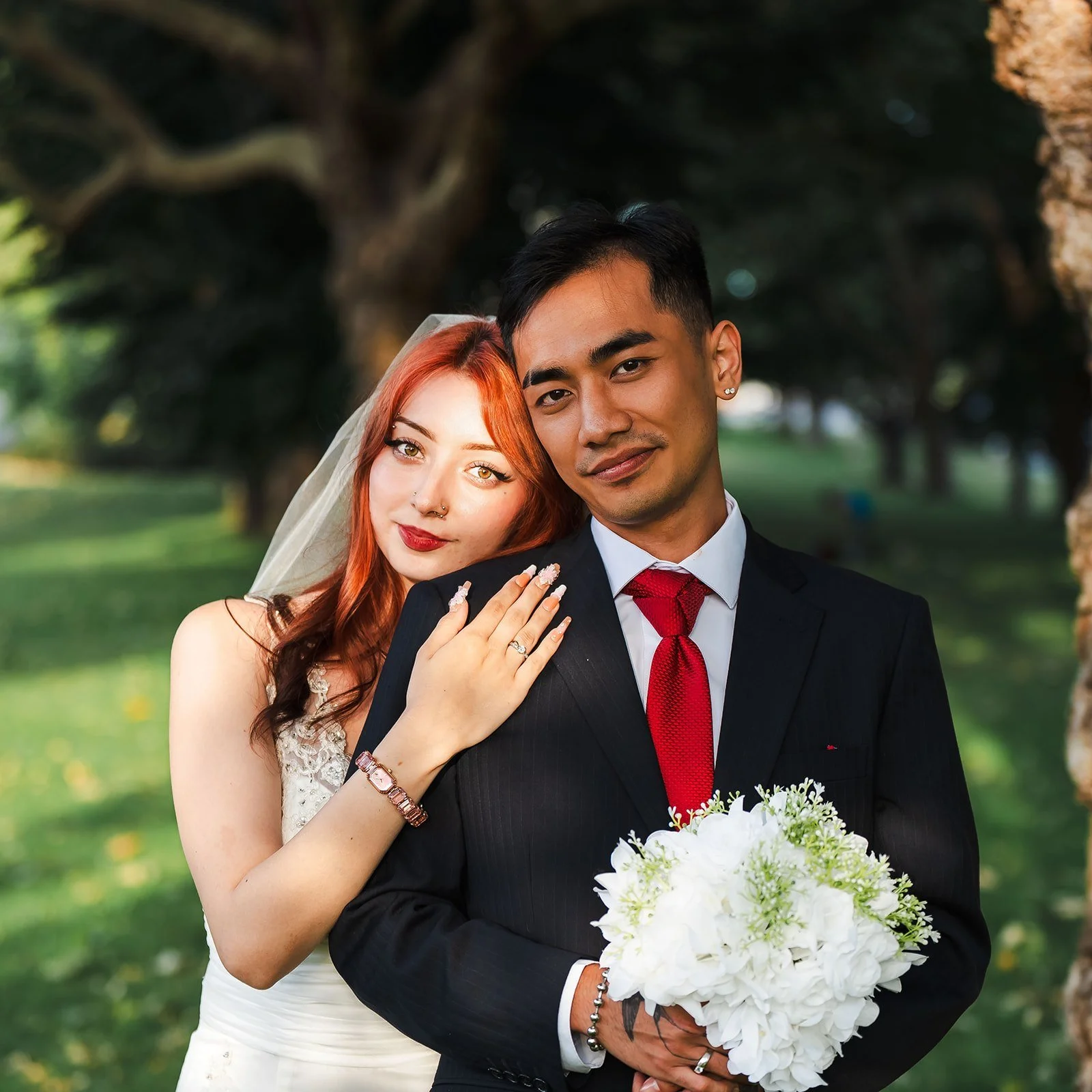 A bride with red hair and a groom with black hair pose outdoors, with the bride resting her hand on the groom's shoulder as she wears a wedding ring, and holding a bouquet of white flowers. They stand in front of trees and greenery.