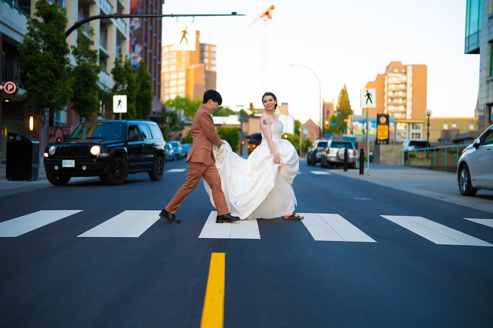 A couple in wedding attire crossing city street on a pedestrian crosswalk, with tall buildings and cars in the background.