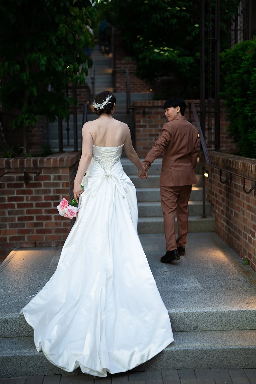 A bride and groom holding hands and walking up outdoor stairs, with the bride in a white wedding dress holding pink flowers, and the groom in a brown suit. The scene appears to be at dusk in a garden setting.