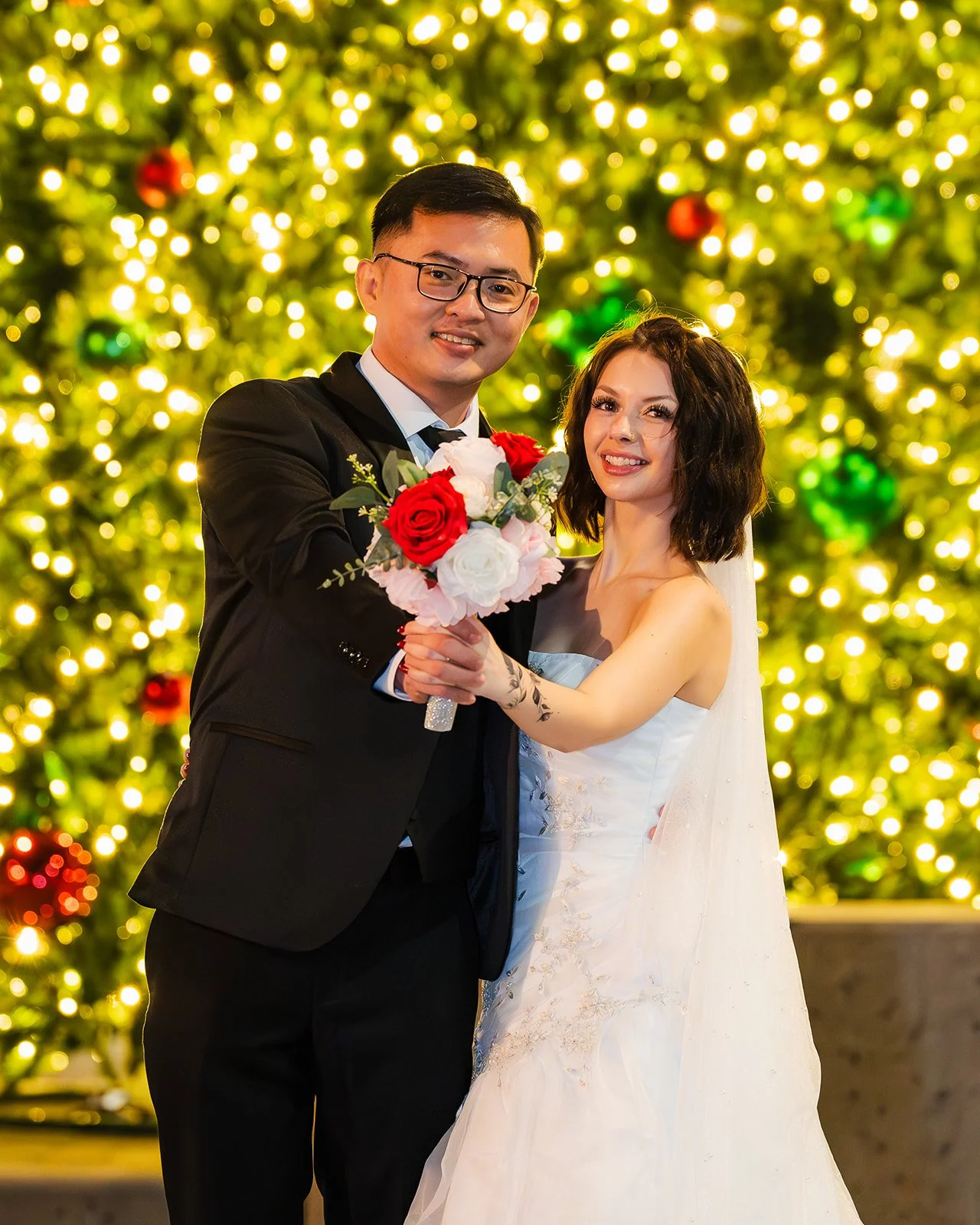A bride and groom holding a bouquet of red and white roses, smiling in front of a decorated Christmas tree with lights and ornaments.