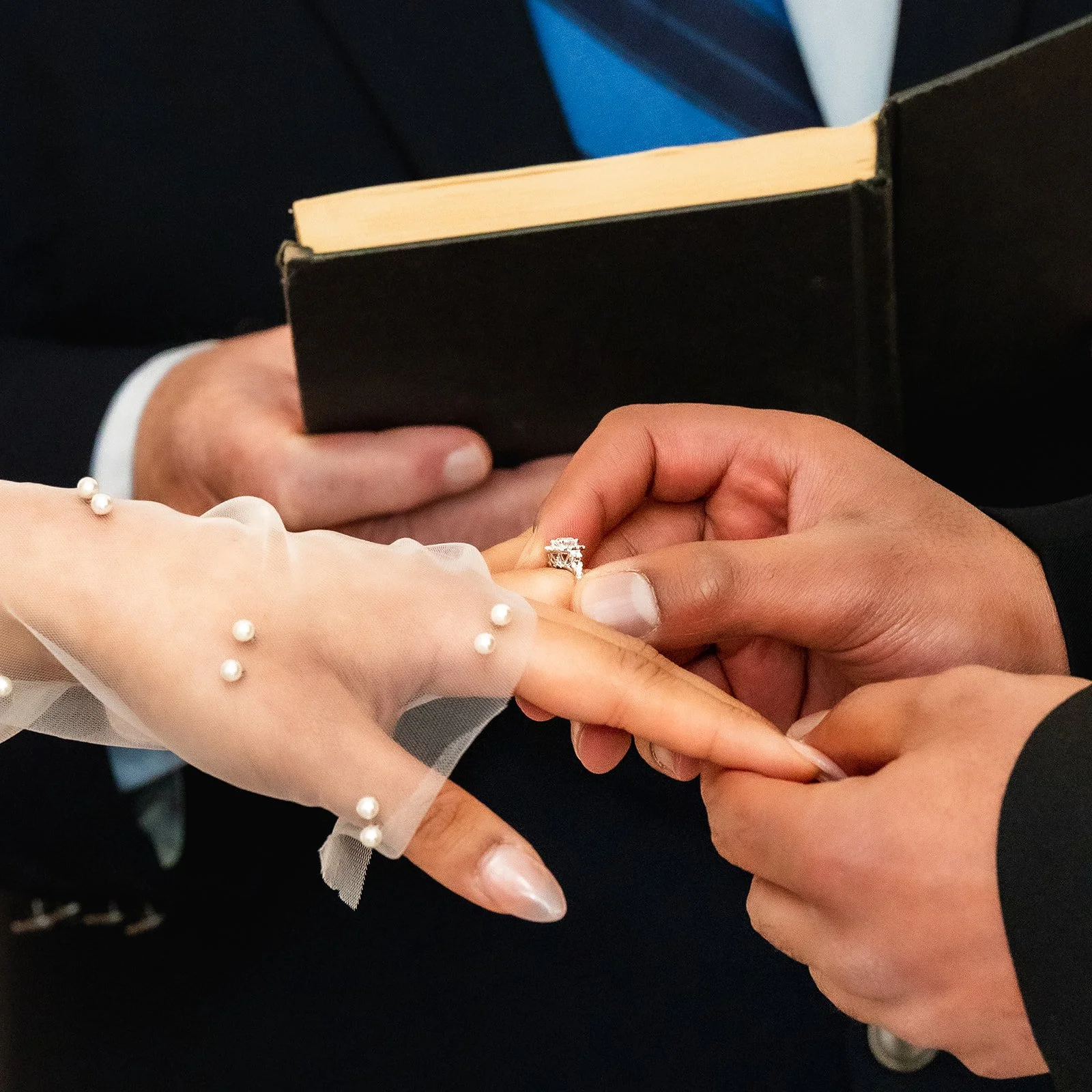 Close-up of a wedding ceremony, showing a bride with a pearl-embellished veil and a groom exchanging rings, with a book in the background.